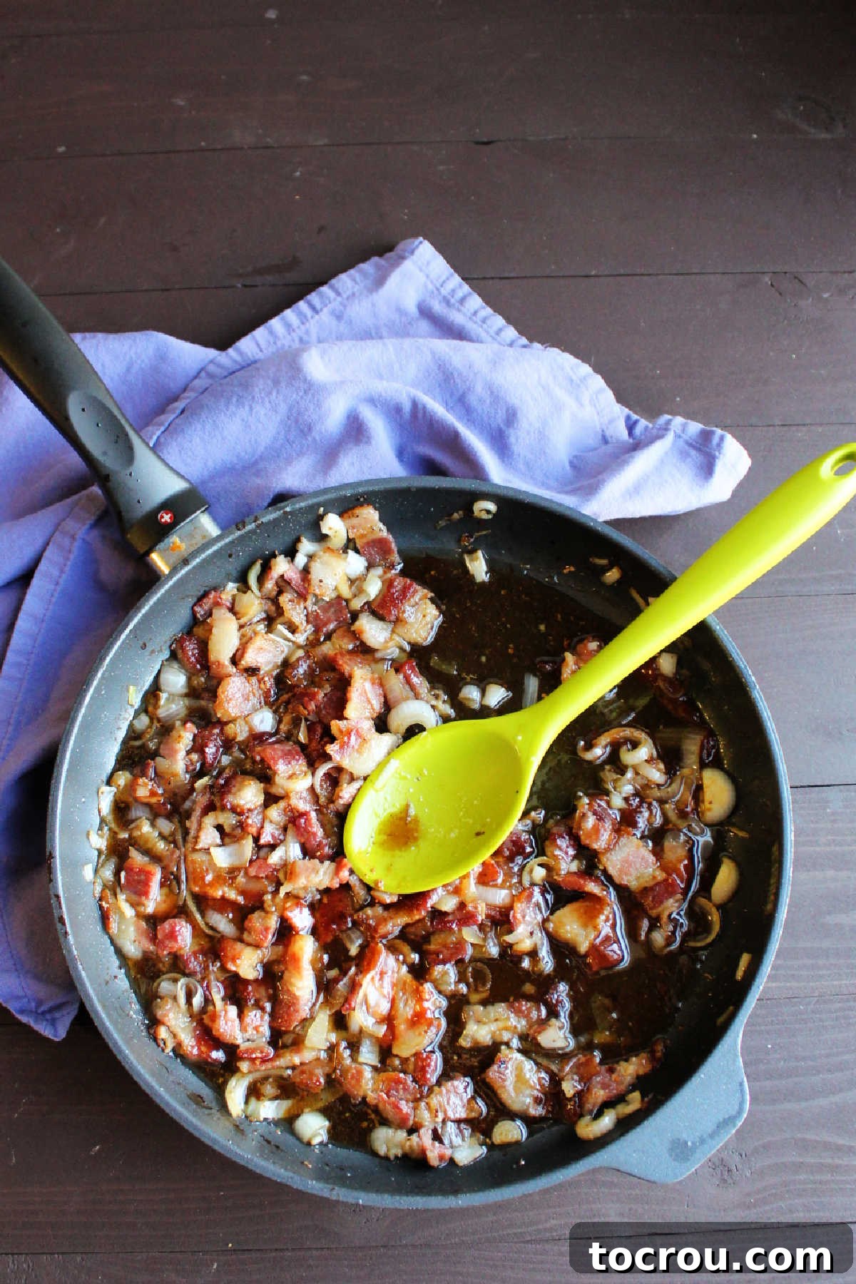 A cast iron skillet filled with warm, bubbling bacon dressing, clearly showing bits of caramelized onion and crispy bacon pieces simmering in the rich liquid.