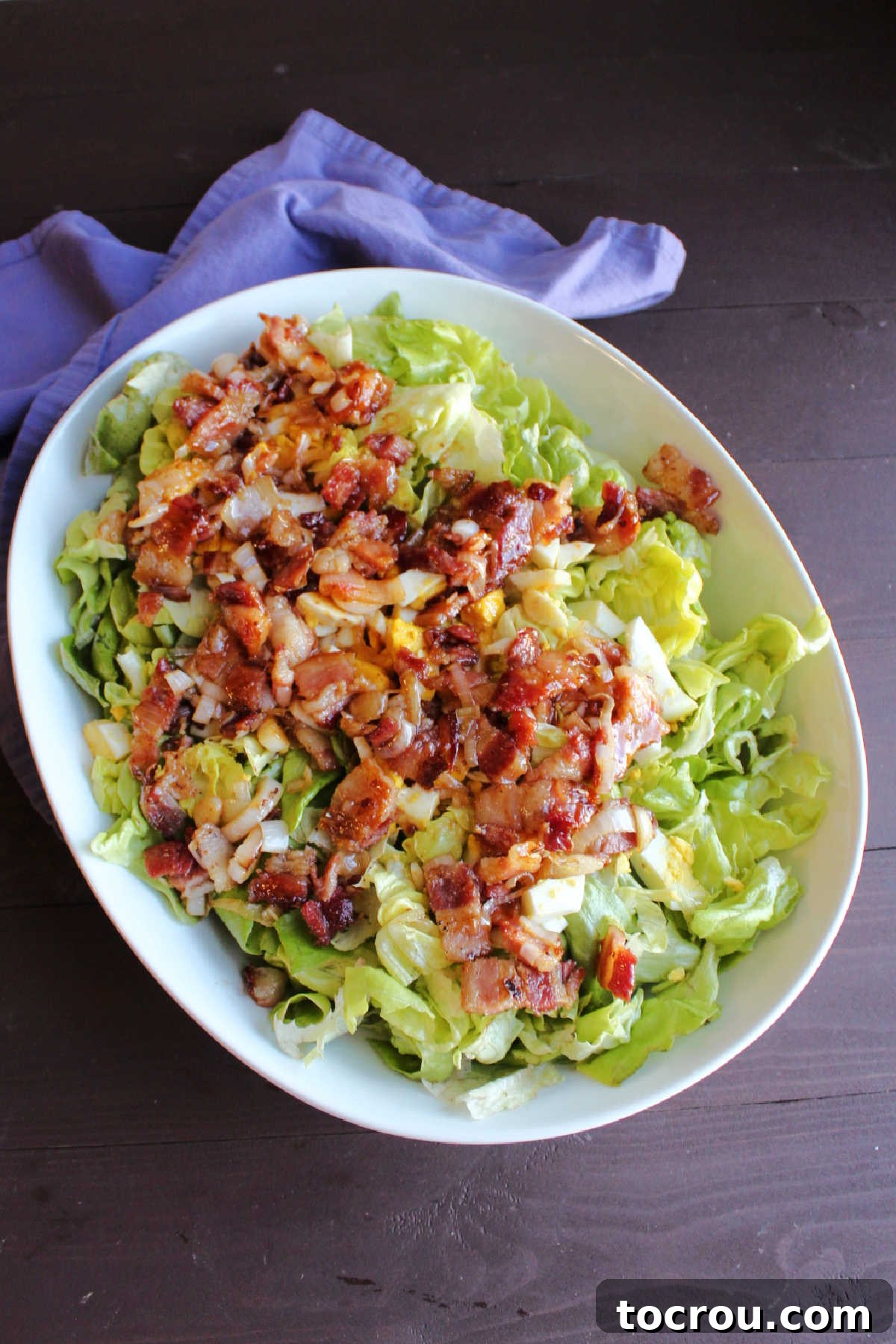 A bowl of fresh lettuce, generously topped with chopped hard-boiled eggs and the warm bacon dressing, poised and ready to be tossed together for serving.