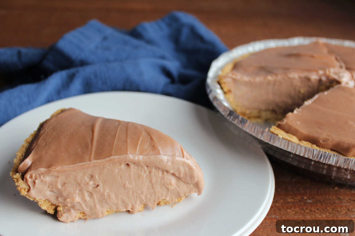 A perfect slice of creamy chocolate no-bake cheesecake pie, adorned with a delicate chocolate curl, sitting on a pristine white plate. The remaining pie, nestled in its foil graham cracker crust, waits patiently in the background, promising more irresistible indulgence.