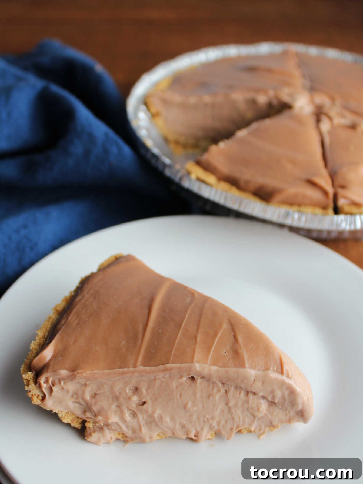 A close-up of a delightful slice of no-bake chocolate cheesecake pie, featuring a golden graham cracker crust and a rich, velvety chocolate cream cheese filling, resting on a dessert plate. The remaining cheesecake is blurred in the background, highlighting the individual serving.