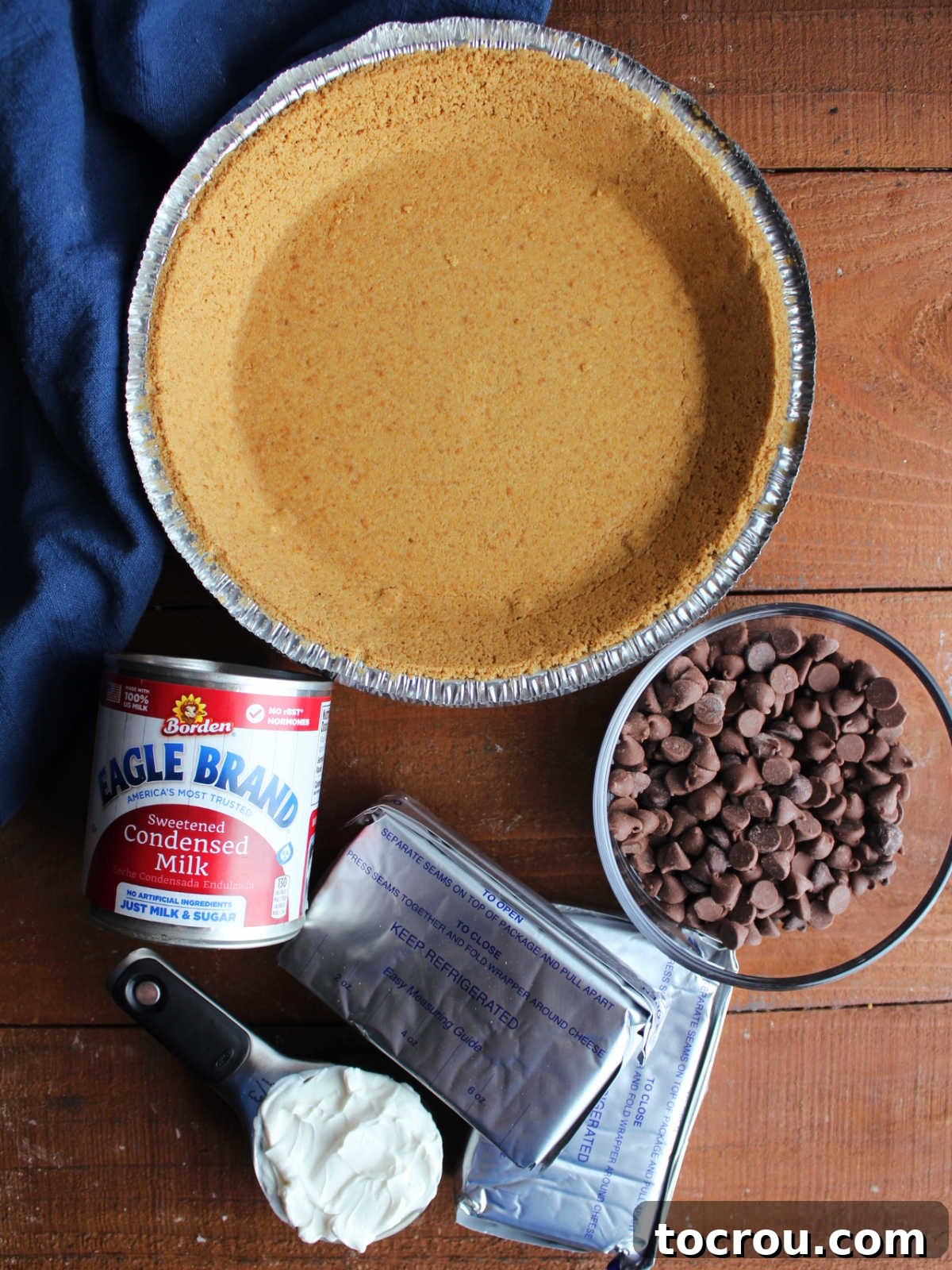A vibrant flat lay shot displaying the key ingredients for a delicious no-bake chocolate cheesecake: a golden graham cracker crust, blocks of softened cream cheese, a can of sweetened condensed milk, a bowl of rich chocolate chips, and a container of creamy sour cream, all ready for preparation.