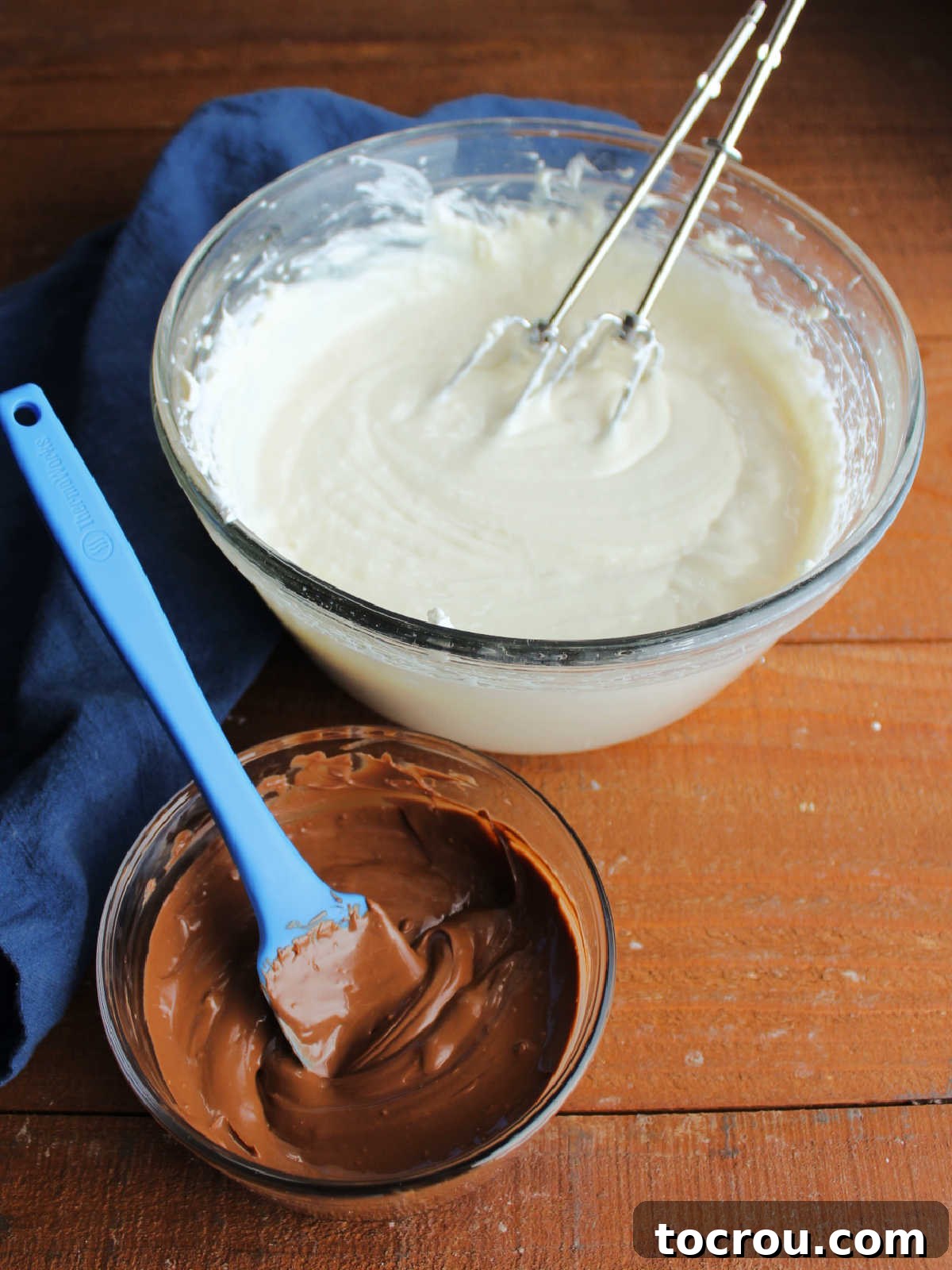 A clear mixing bowl filled with the creamy no-bake cheesecake base, light in color and smooth in texture. Adjacent to it, a smaller bowl holds a pool of glossy, melted chocolate, poised and ready to be incorporated into the mixture, creating the perfect chocolate cheesecake filling.