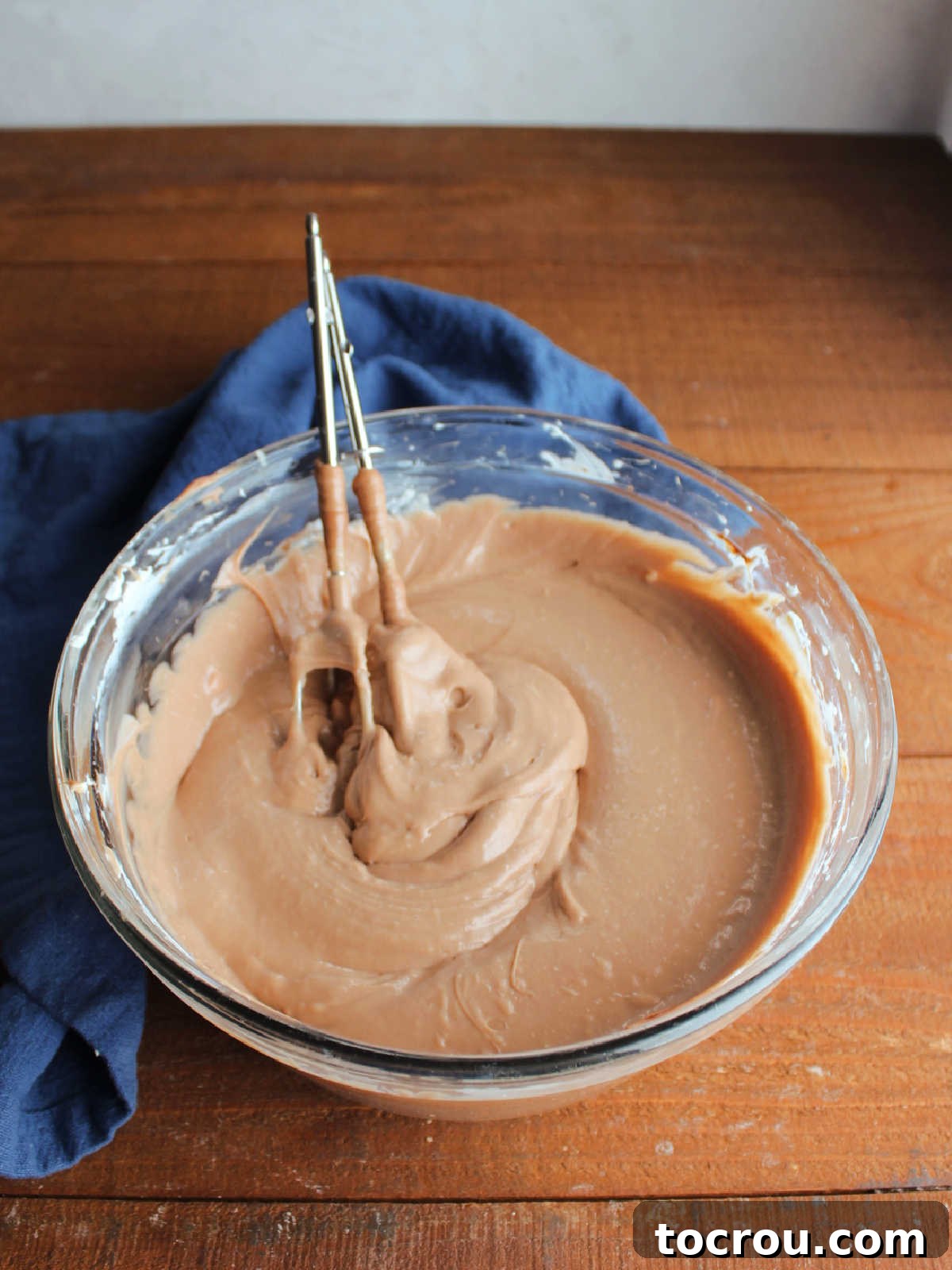 A clear glass mixing bowl contains the rich, dark chocolate no-bake cheesecake mixture, perfectly blended and ready for chilling. A handheld mixer, with beaters still coated in the creamy filling, rests inside the bowl, highlighting the effortless preparation.