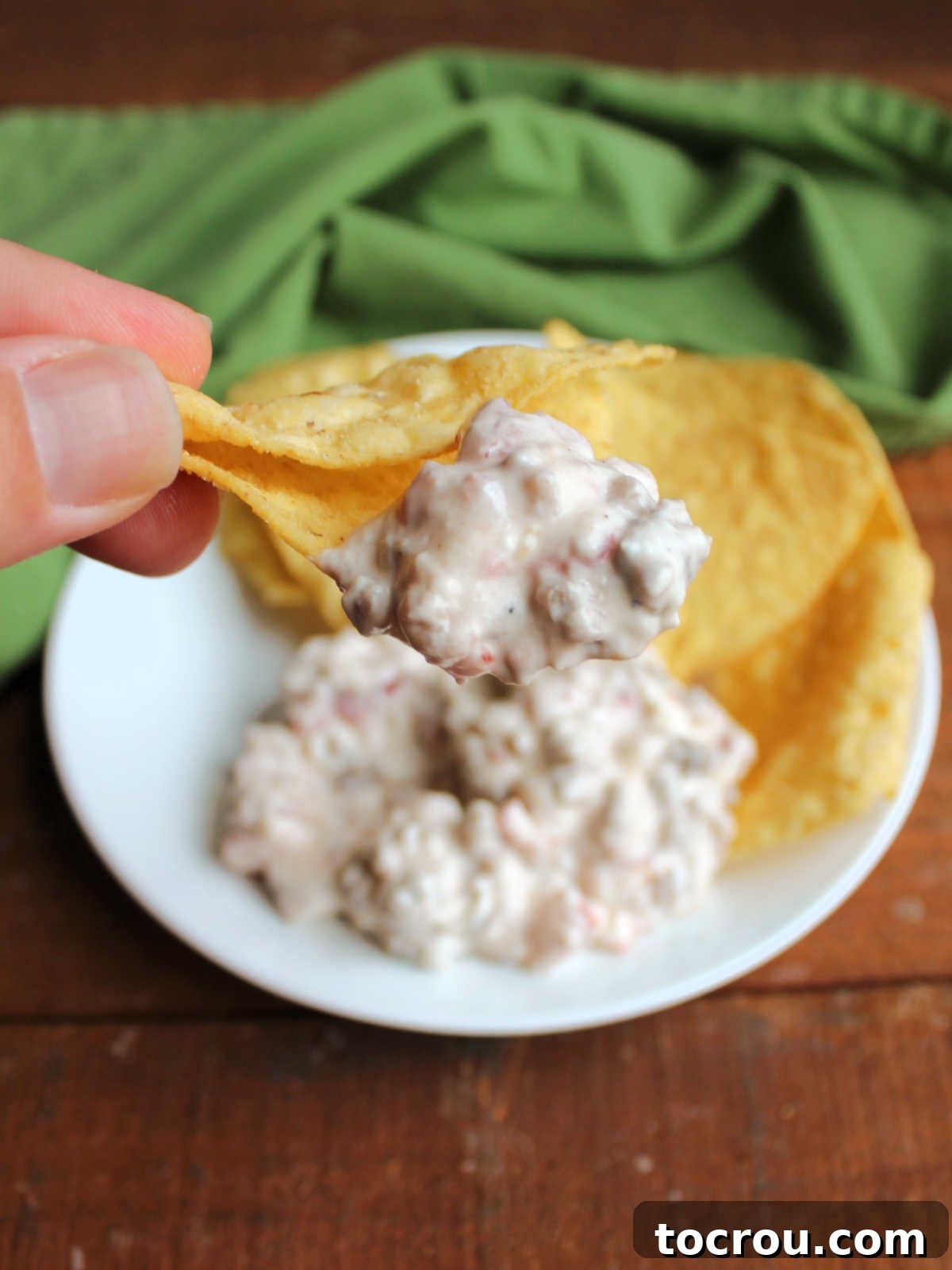 Hand holding a tortilla chip with creamy sausage and Rotel dip on it, ready to eat.