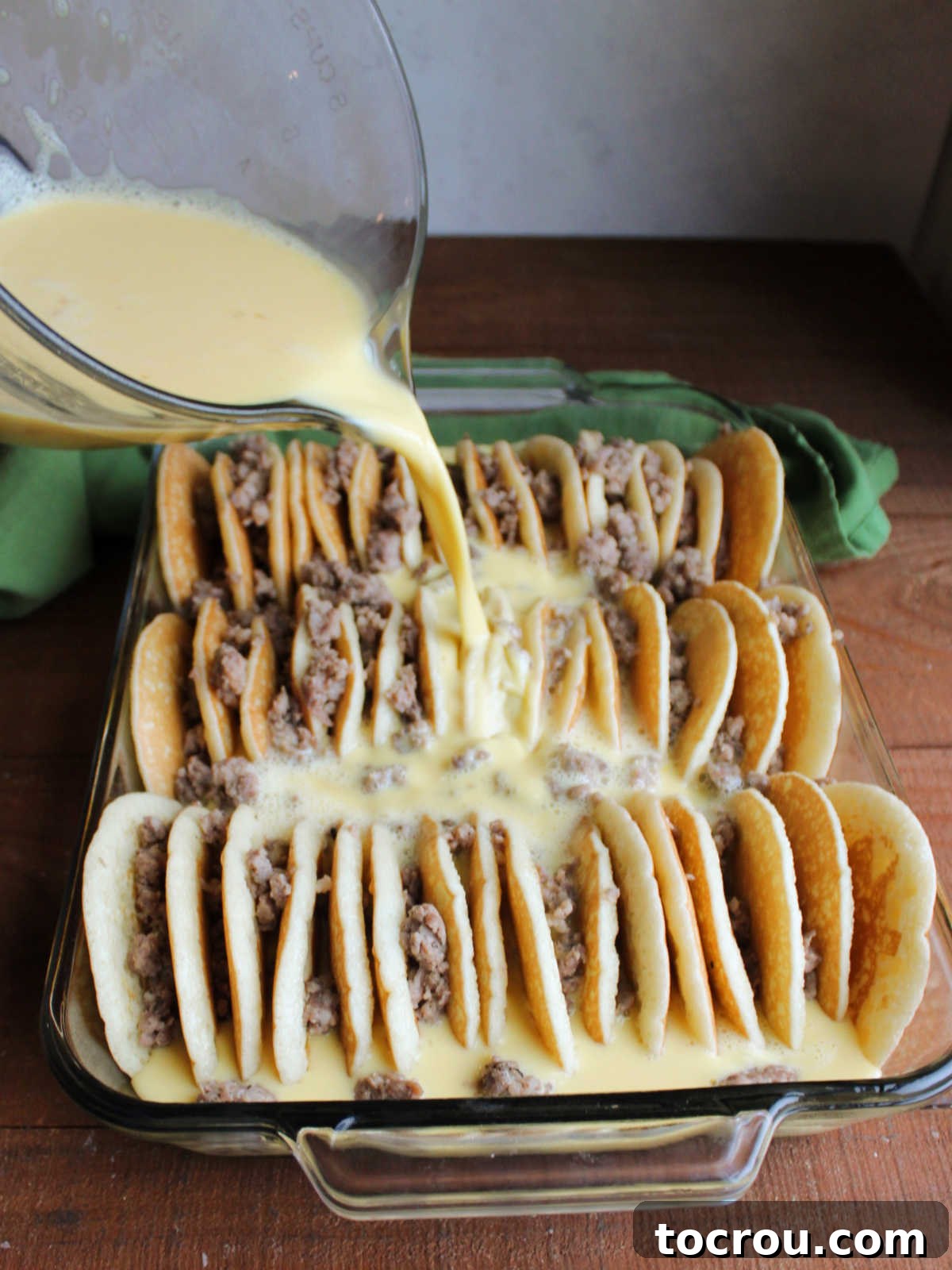 Casserole dish filled with pancake halves that are arranged with the cut side down and curved edges up with sausage between the pancakes making them almost look like tacos with egg and milk mixture being poured over them.