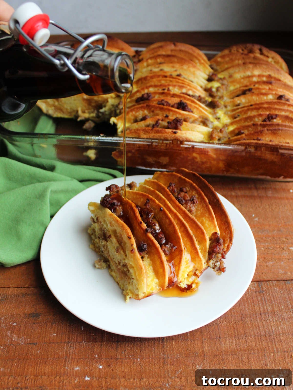 Pouring maple syrup over a piece of pancake and sausage bake on a small plate. 