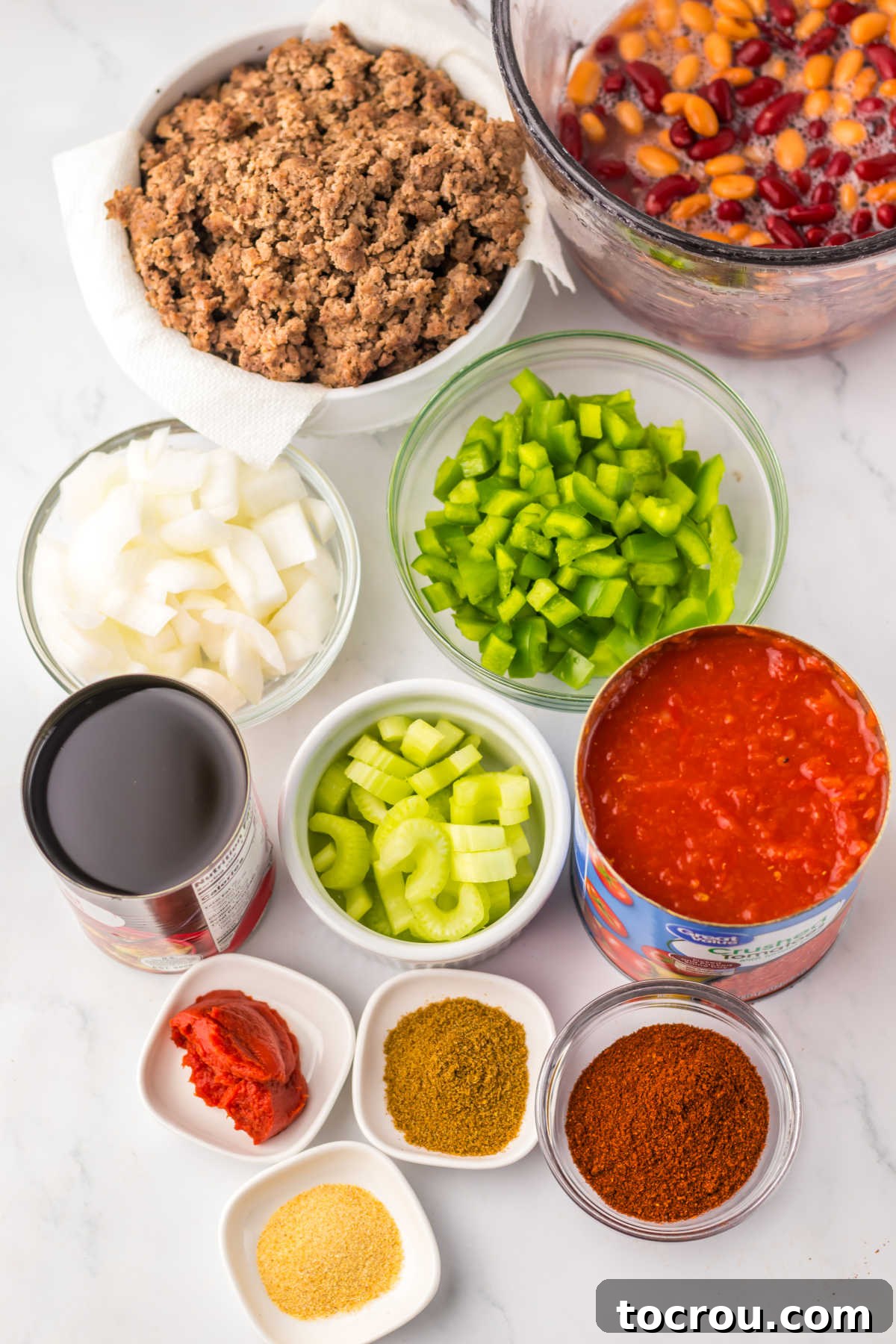 Ingredients for slow cooker chili including cooked ground beef, various canned beans, fresh green pepper, onion, celery, canned tomatoes, and a selection of seasonings.