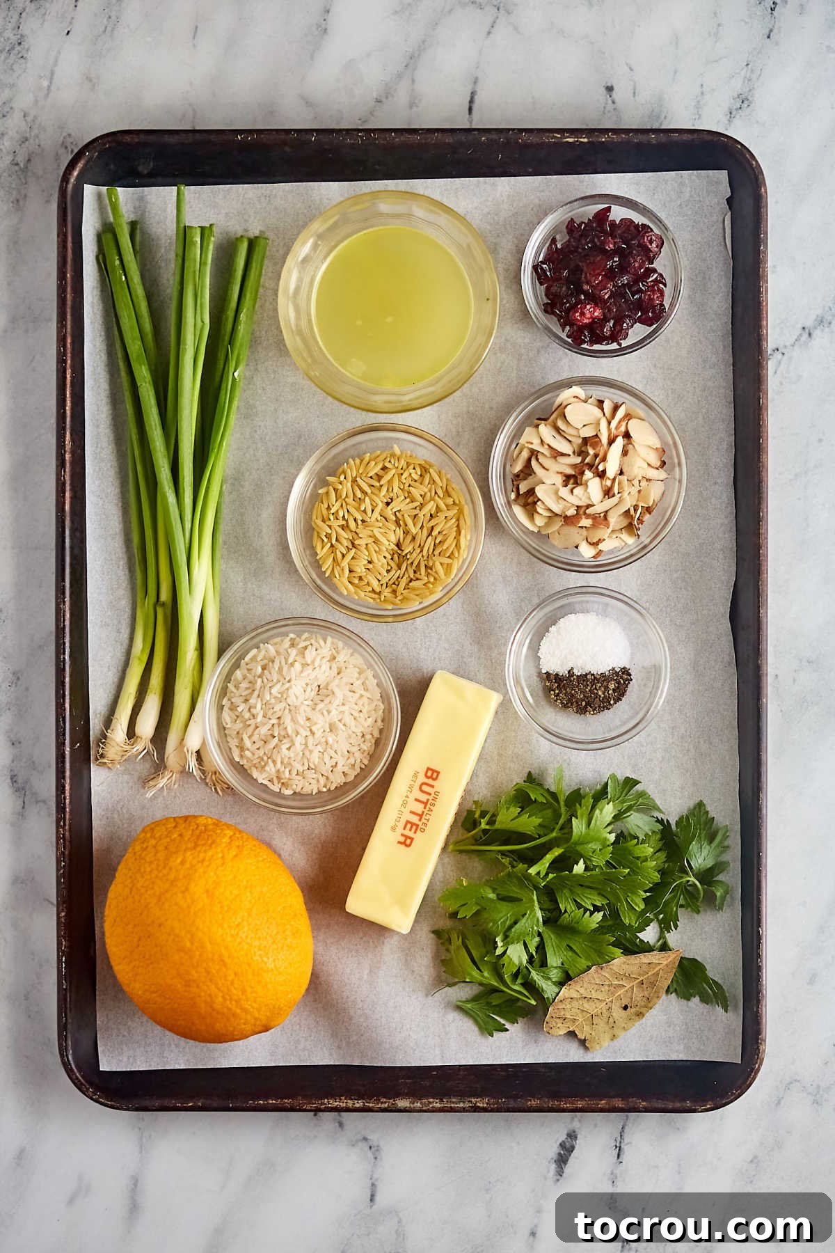 Fresh Ingredients for Rice Pilaf A selection of fresh ingredients laid out for homemade Orange Cranberry Rice Pilaf, including long-grain rice, orzo, butter, chicken broth, dried cranberries, slivered almonds, salt, pepper, vibrant green onions, a large navel orange, fresh parsley, and a bay leaf.