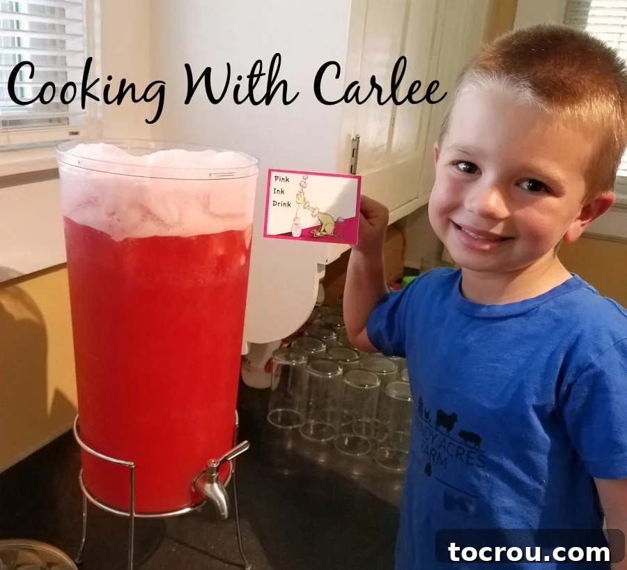 A young boy holds a 'pink ink drink' sign next to a large beverage dispenser filled with vibrant pink punch.