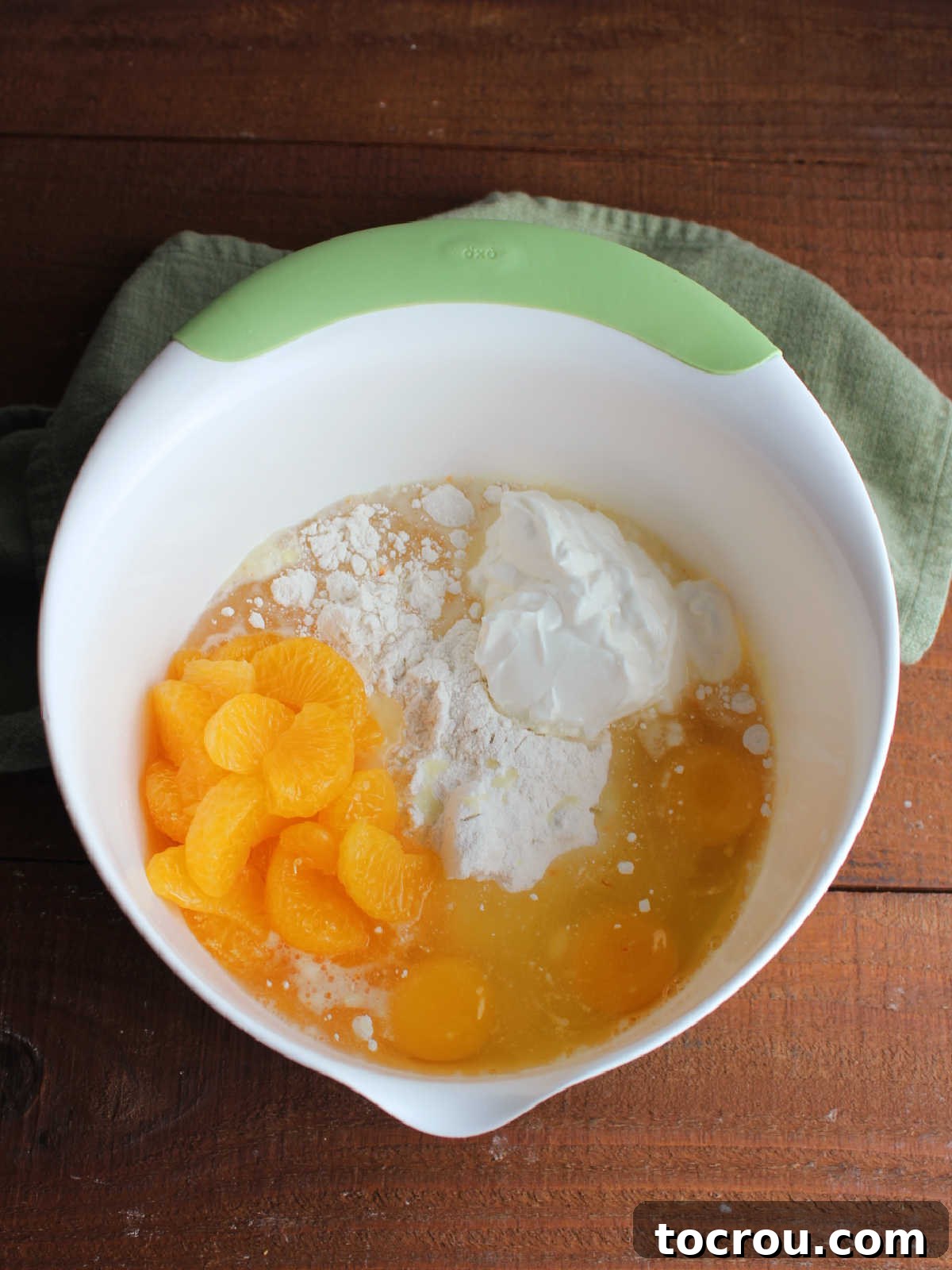 Cake Base Ingredients A large mixing bowl containing a yellow cake mix, a can of mandarin oranges (undrained), and sour cream, ready to be combined for the Ambrosia Poke Cake batter.