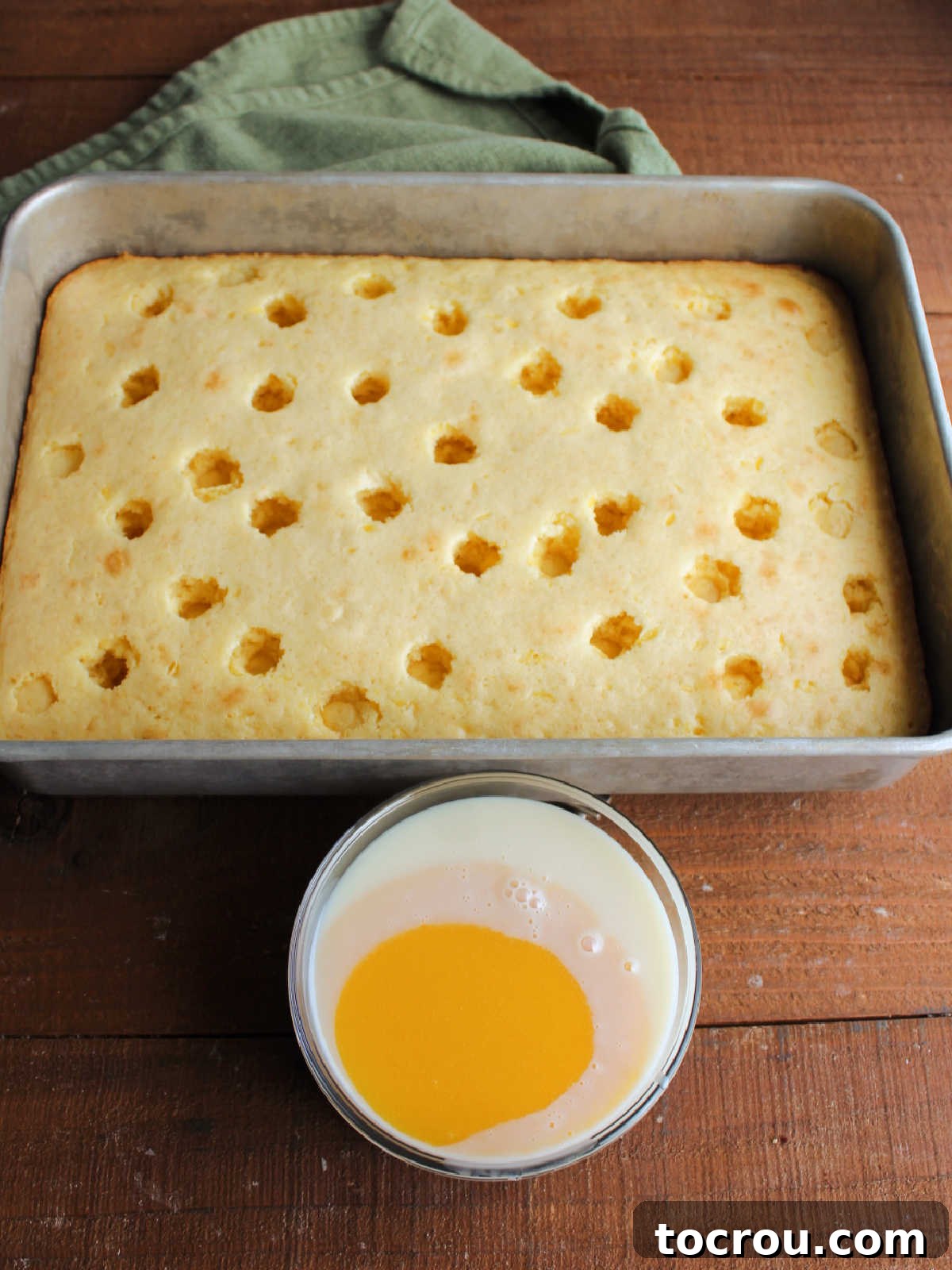 Poke Cake Ready for Soak A freshly baked cake with holes poked across its surface, ready for soaking, sits beside a small bowl containing the golden mixture of sweetened condensed milk and orange juice concentrate.