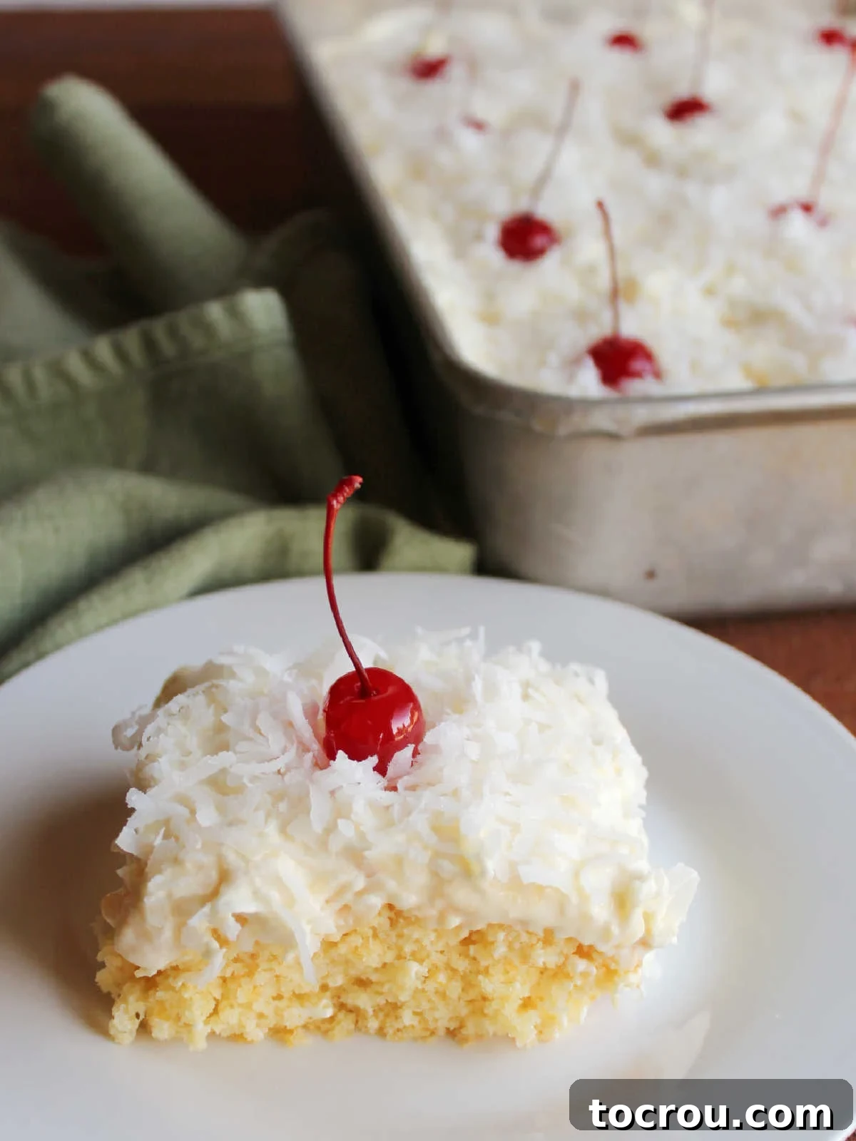 Ambrosia Poke Cake Plated A beautifully presented slice of Ambrosia Poke Cake, featuring the vibrant orange condensed milk soak and the exquisite pineapple, coconut, and cherry topping, on a plate with the remaining cake visible in the background.