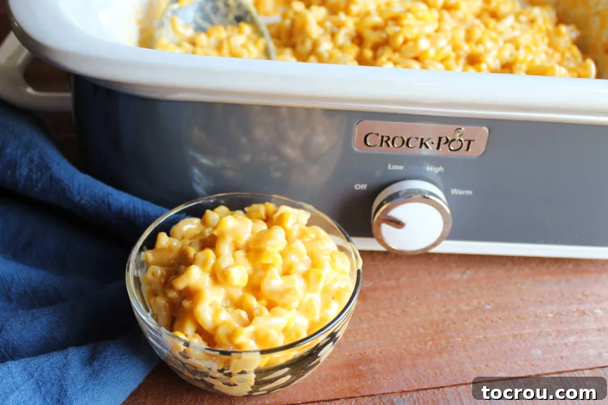 A bowl of creamy macaroni and cheese with corn, served in front of a slow cooker with the remaining casserole inside. The texture is smooth and inviting, showcasing perfectly cooked elbow macaroni and golden corn kernels.