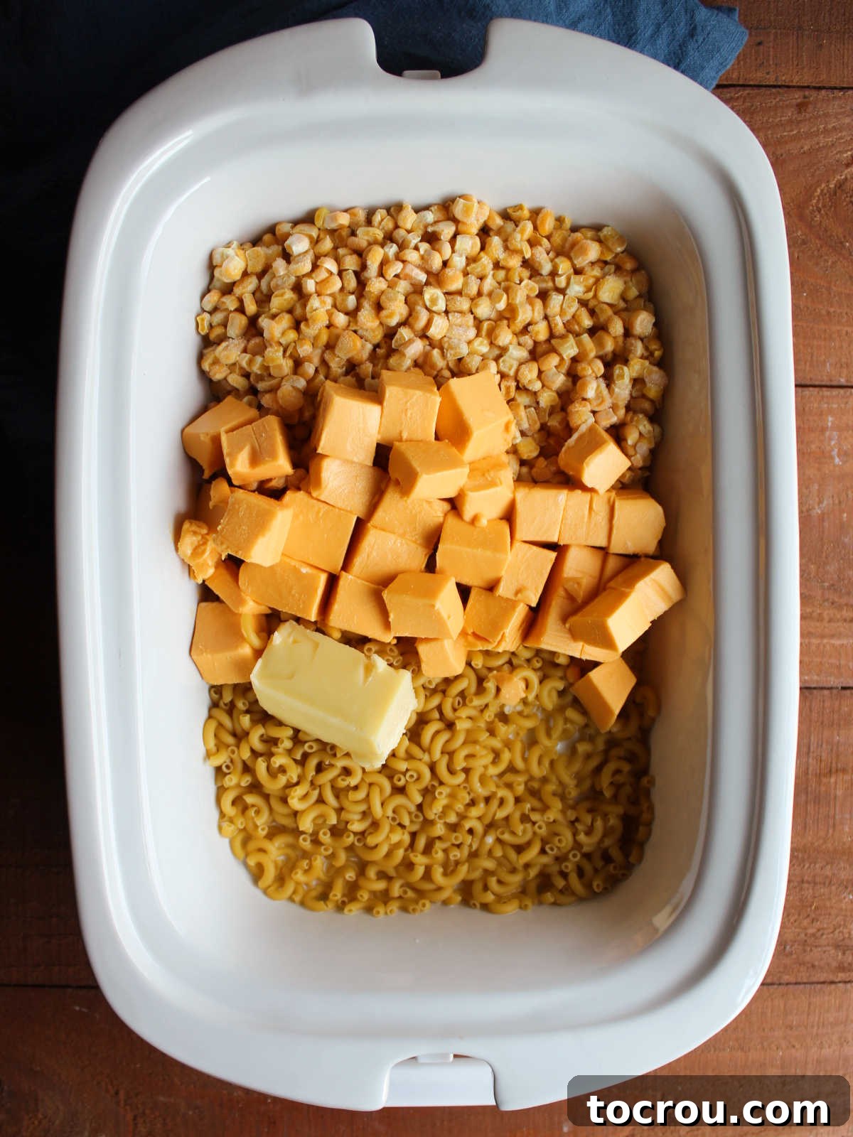 A slow cooker filled with uncooked macaroni noodles, frozen corn, cubed Velveeta cheese, pats of butter, milk, and water, ready for cooking. The ingredients are neatly arranged, showcasing the simplicity of the preparation.