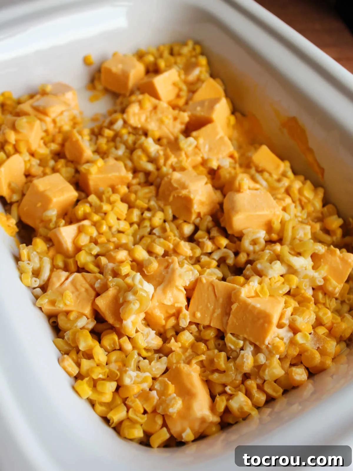 A close-up view of the macaroni and corn casserole in the slow cooker during the initial cooking phase. The cheese is beginning to melt, but the macaroni noodles are still firm, illustrating the progress of the slow cooking process.