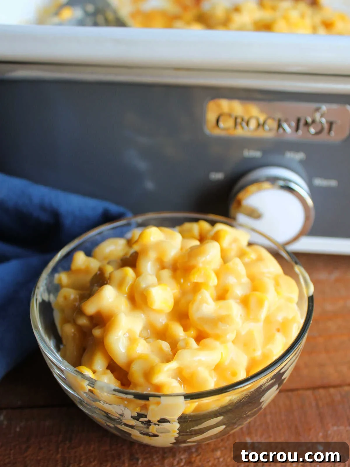 A serving of corn macaroni and cheese in a glass bowl, positioned in front of the slow cooker it was cooked in. The dish exhibits a smooth, creamy cheese texture with perfectly cooked elbow macaroni noodles and corn kernels visible.