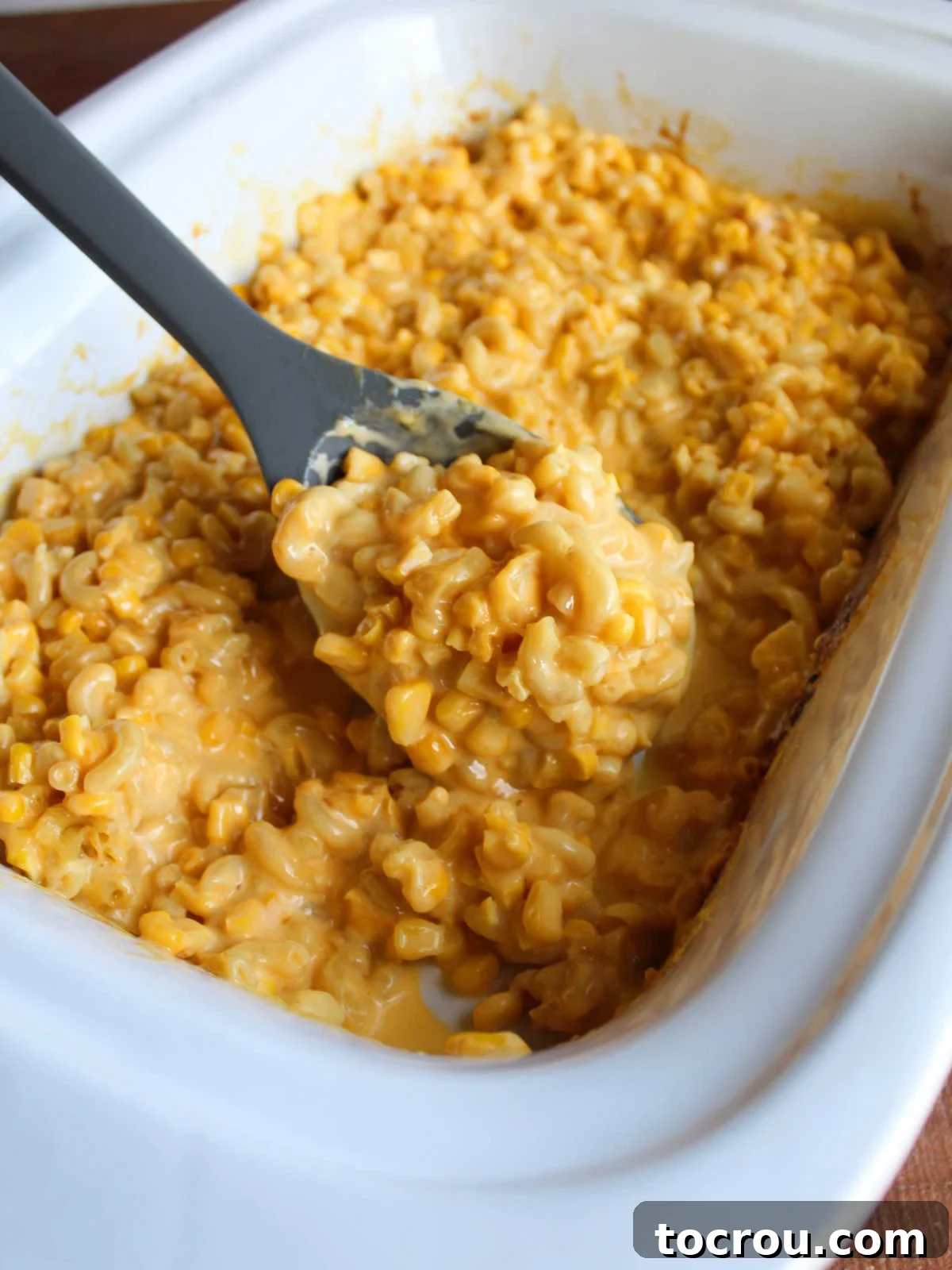 A serving spoon lifting a generous scoop of the creamy corn macaroni and cheese from the slow cooker, ready to be served. The texture is luscious, and the steam suggests it's fresh and hot.