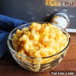Small glass bowl filled with cheesy macaroni and corn casserole in front of the crockpot it was cooked in.