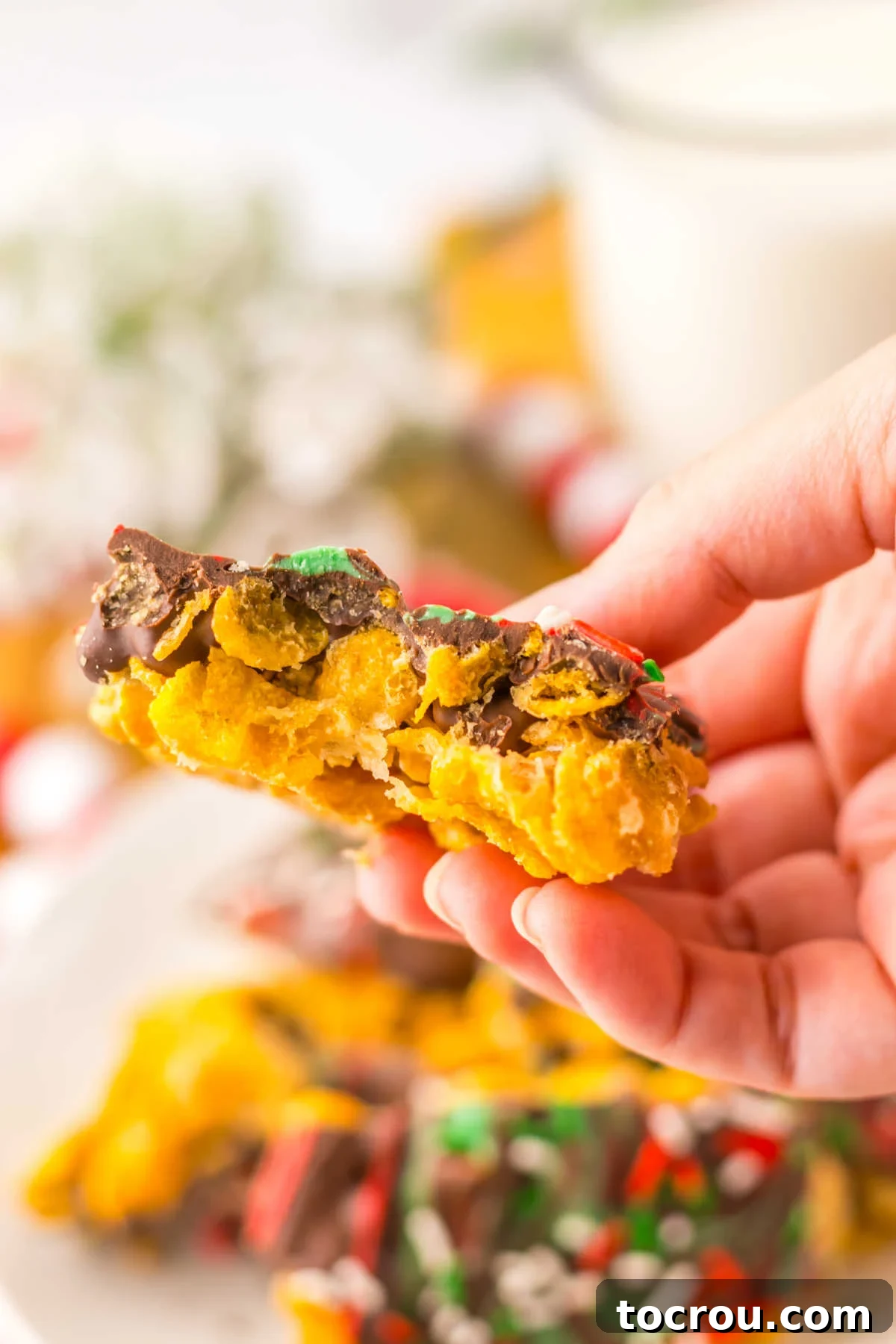 A hand holds a piece of cornflake brittle, highlighting its inviting, crunchy, caramelized texture and the delicate layer of chocolate on top, ready to be enjoyed.