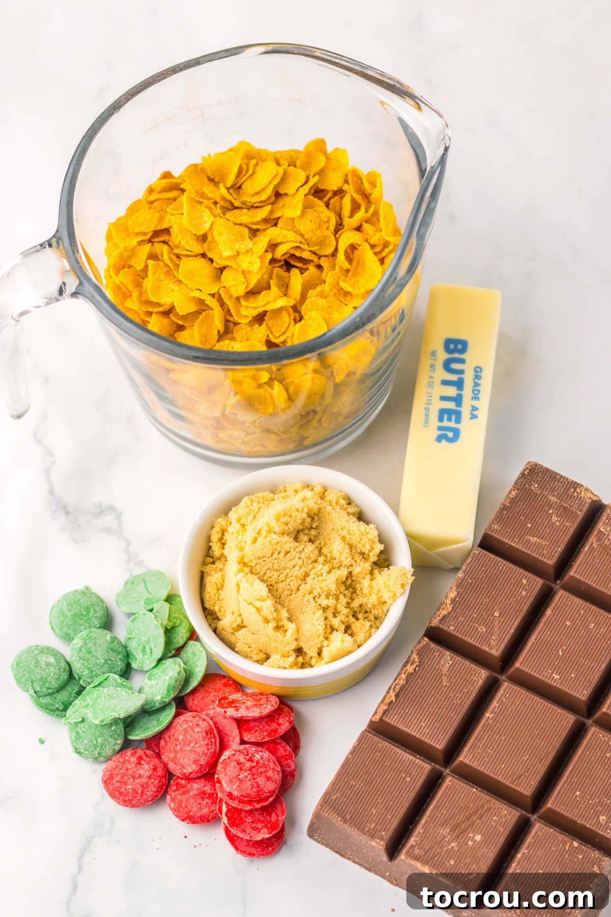 A vibrant collection of ingredients for festive cornflake brittle, including golden cornflakes, creamy butter, rich brown sugar, dark chocolate, and brightly colored red and green candy melts, arranged and ready for preparation.