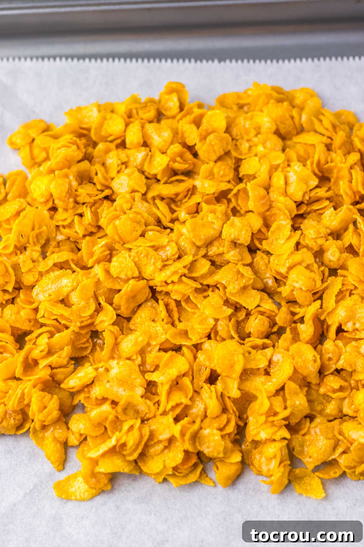 A baking pan lined with parchment paper holds a spread of cornflakes generously coated in a shimmering butter and brown sugar mixture, ready for baking.