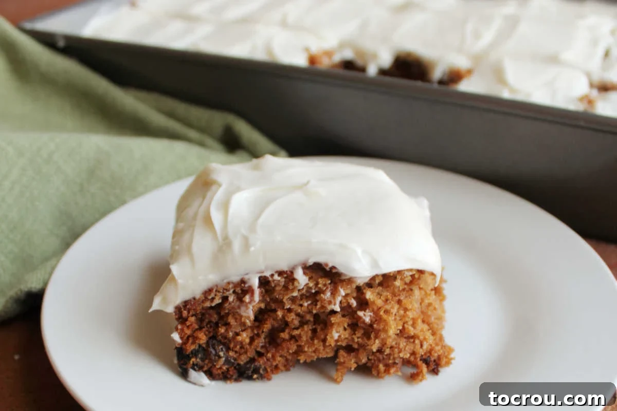 A generous piece of moist, spiced applesauce cake with raisins and a luscious cream cheese frosting, artfully placed in front of a 9x13-inch baking pan containing the rest of the delectable cake.
