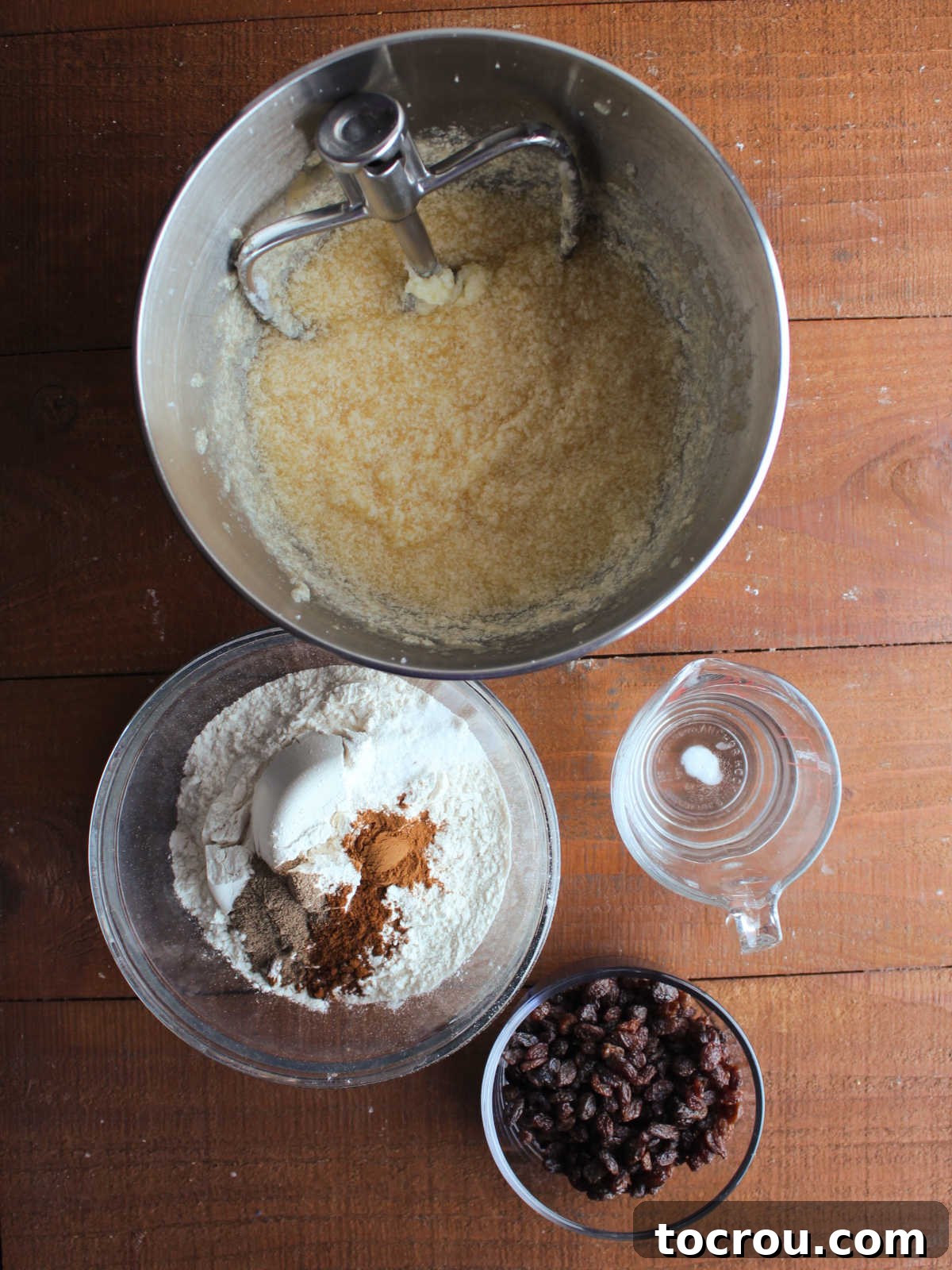 The initial stage of making applesauce cake batter, showing a mixer bowl filled with creamed butter, sugar, and egg mixed with applesauce. Adjacent are a mixing bowl with dry ingredients, a small bowl of raisins, and a measuring cup holding hot water and baking soda, all ready for combination.