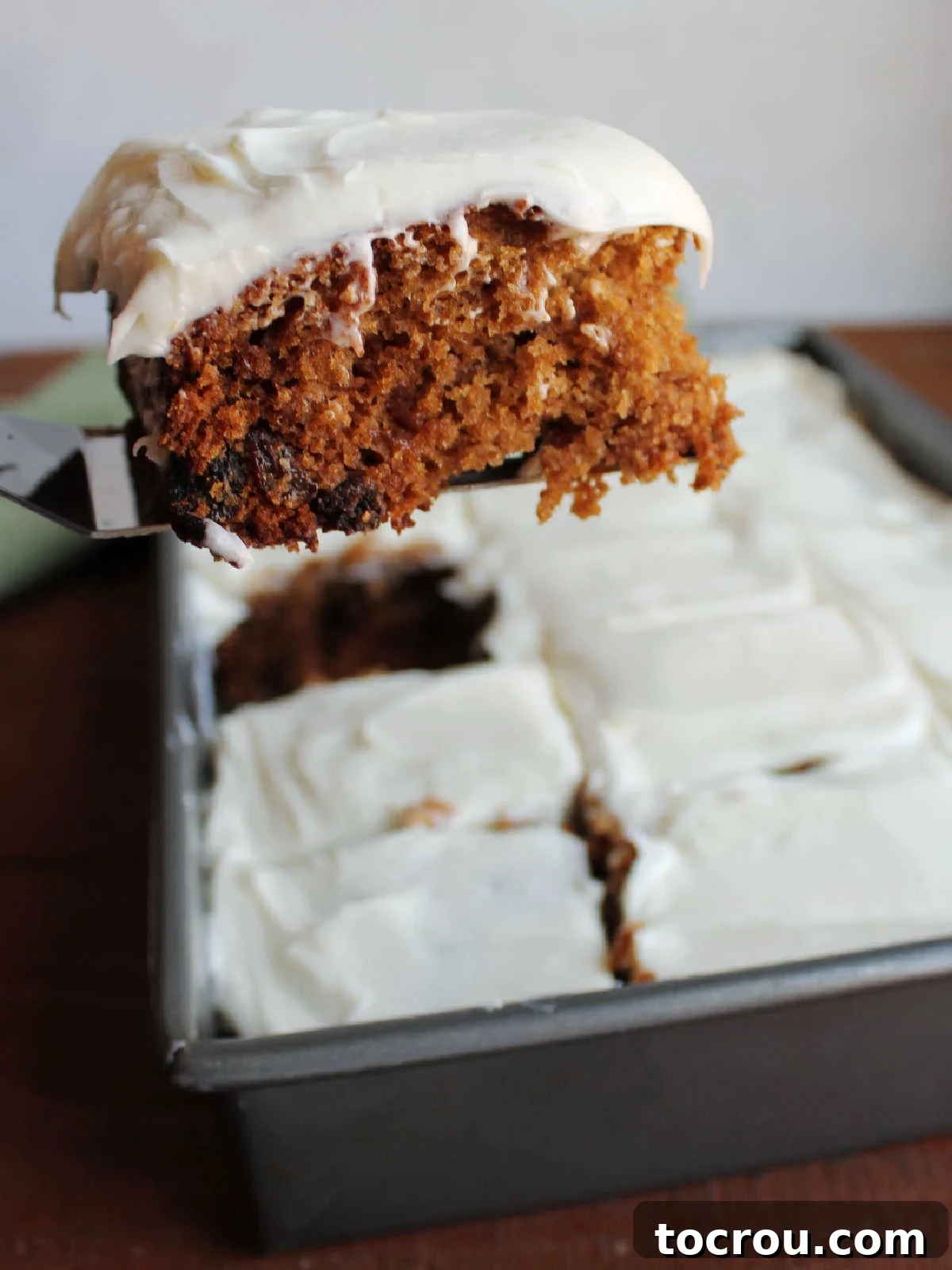 A spatula gently lifting a slice of applesauce spice cake, revealing its moist texture and deep brown color, studded with raisins and topped with a swirl of cream cheese frosting.