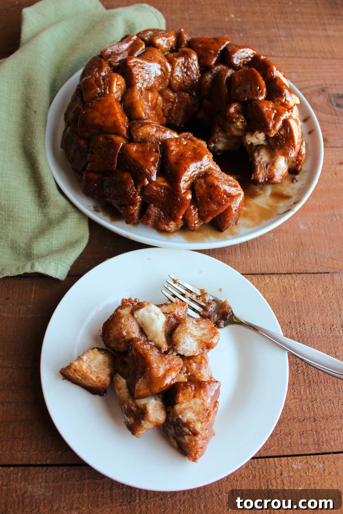 Serving of monkey bread on a small plate with a fork with the remaining brown sugar and cinnamon monkey bread in the background. 