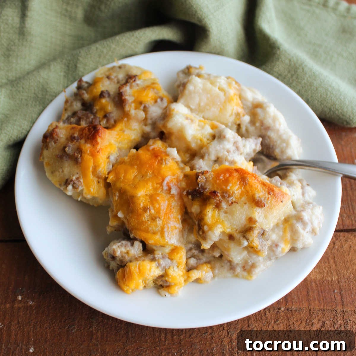 Biscuits and Gravy Bubble Up Bake Small plate piled high with fluffy biscuit pieces soaked in creamy sausage gravy and topped with golden-brown melted cheddar cheese, with a fork ready to scoop.