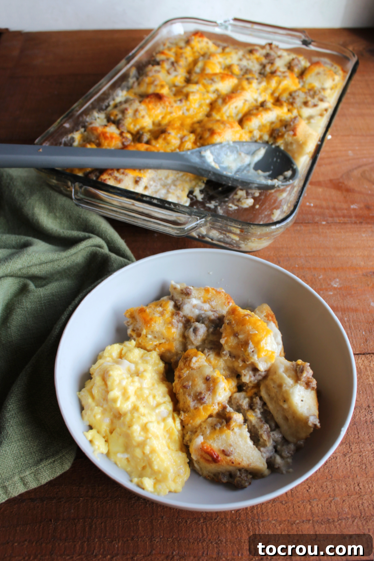 Served Biscuits and Gravy Casserole with Eggs A generous serving of Biscuits and Gravy Bubble Up Bake in a shallow bowl, accompanied by soft scrambled eggs, with the remaining golden-brown casserole in the background.