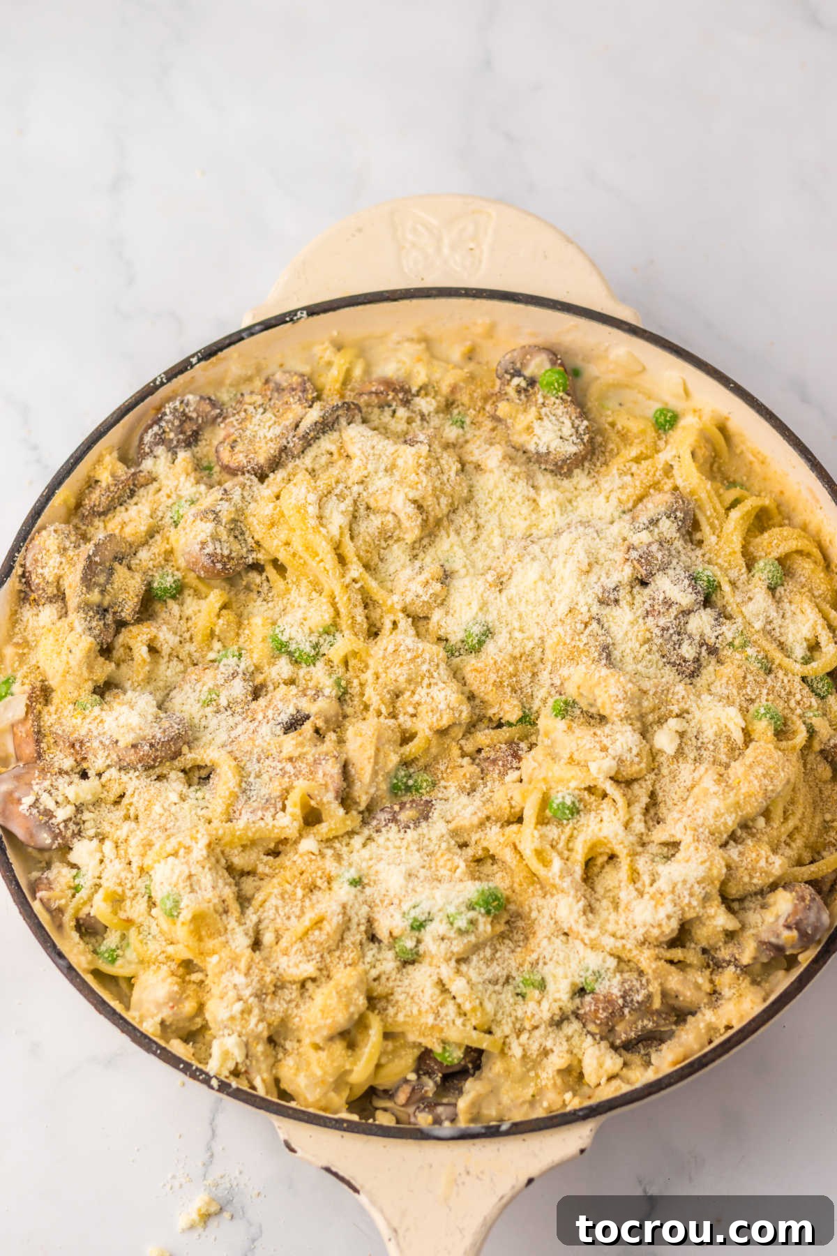 Deep enameled cast iron skillet filled with pasta in creamy sauce with turkey, peas, and mushrooms topped with bread crumbs and cheese ready to go in the oven.