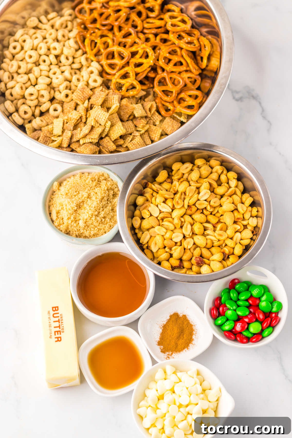 A collection of ingredients for sweet Chex mix, including Chex cereal, Cheerios, pretzels, peanuts, butter, maple syrup, brown sugar, cinnamon, vanilla extract, M&Ms, and white chocolate chips, arranged for slow cooker preparation.