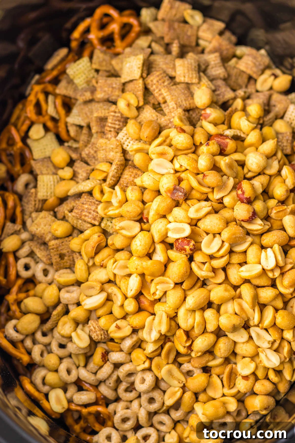 A slow cooker bowl filled with a base mixture of peanuts, Cheerios, Chex cereal, and mini pretzels, ready for the sweet maple cinnamon coating.