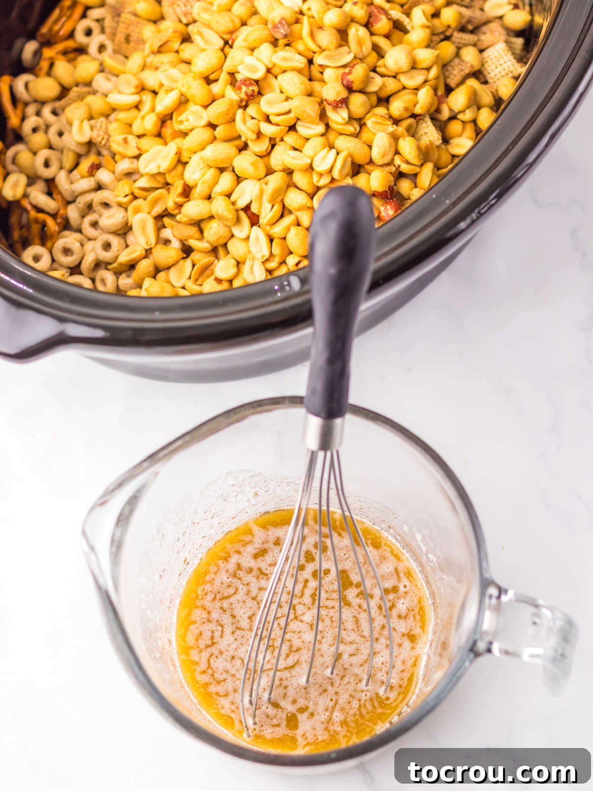 A glass measuring cup containing a rich, melted mixture of butter, brown sugar, maple syrup, vanilla, and cinnamon, prepared to be poured over the cereal blend in the slow cooker.