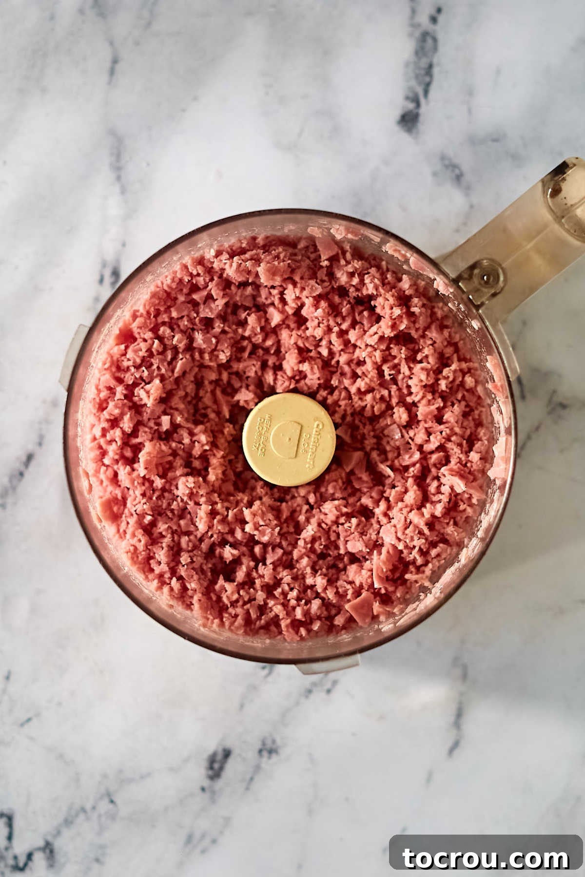 Close-up image of finely ground ham in a food processor, showing distinct, small crumbles rather than a pasty texture, perfectly prepared for ham loaf.