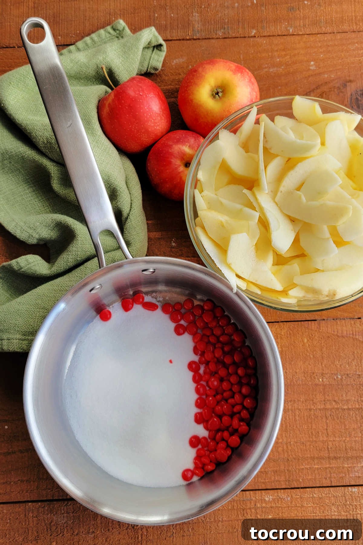 Saucepan with sugar and red hots next to a bowl of thinly sliced peeled apples.