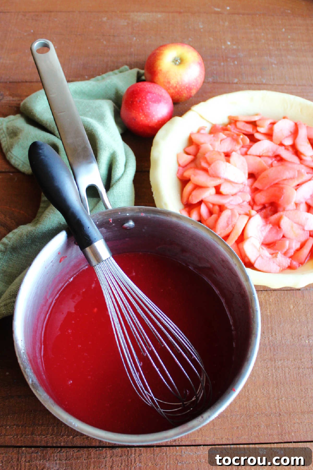 Saucepan of thickened red hot and sugar syrup next to a pie plate with the bottom crust and the partially cooked apple slices.