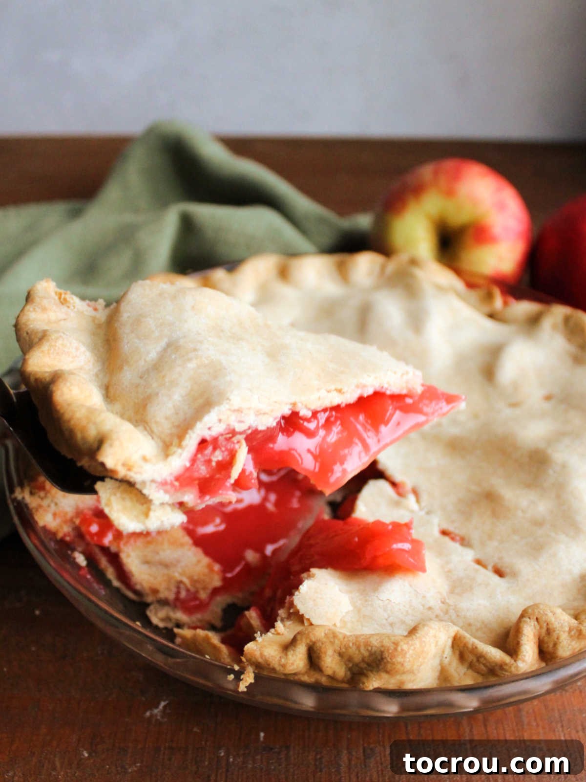 Lifting a sliced of red hot cinnamon apple pie out of the pie plate showing red apple filling.