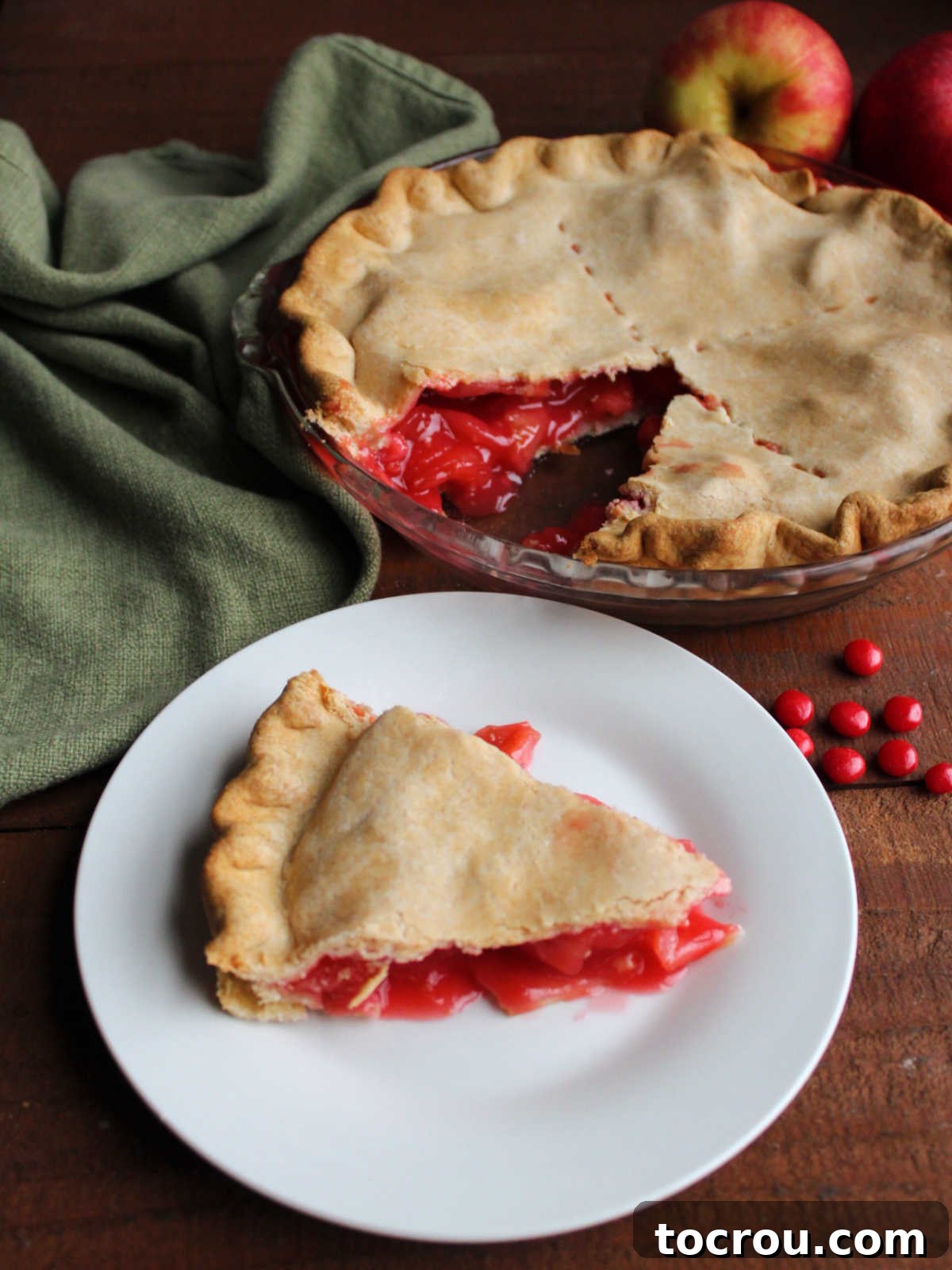 Slice of red hot apple pie on plate with remaining pie in the background.
