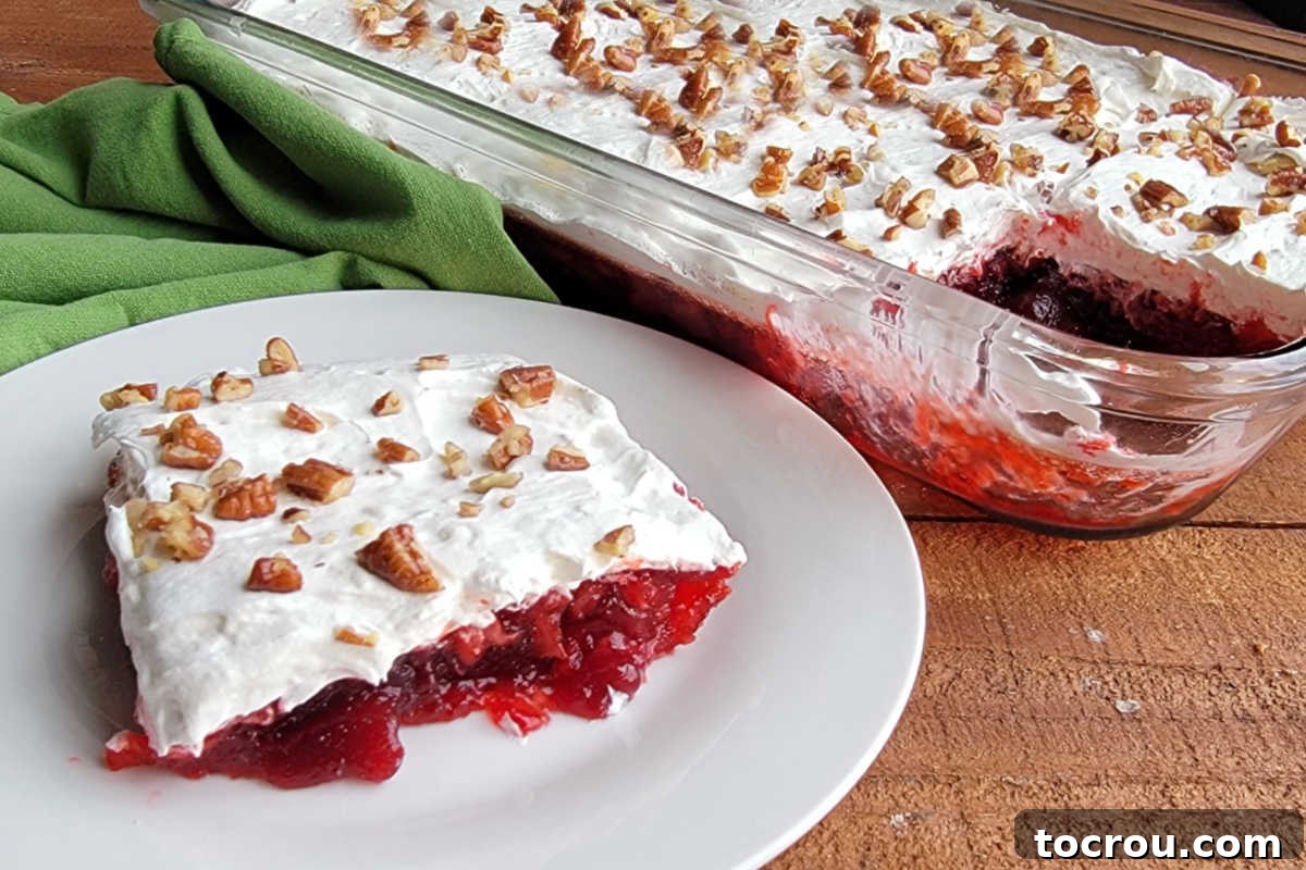 A square piece of frosted cranberry Jello salad, showcasing a bright red Jello base filled with fruit, topped with a fluffy white cream cheese layer and sprinkled with toasted pecans, resting on a white plate.