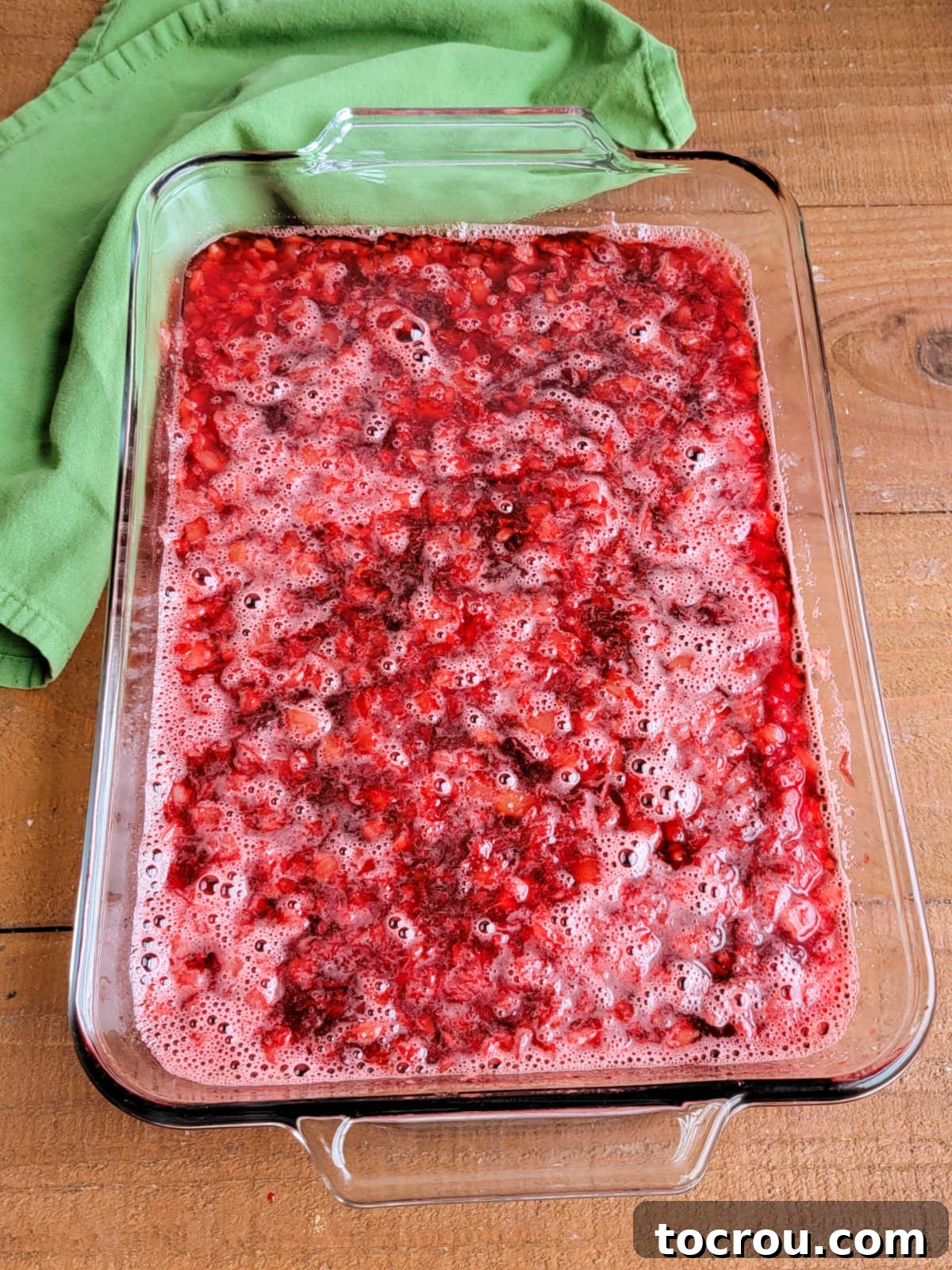 A clear 9x13-inch glass baking dish filled with the bright red Jello mixture, showcasing pieces of crushed pineapple and cranberry sauce, with the subtle fizz of Coca-Cola visible.