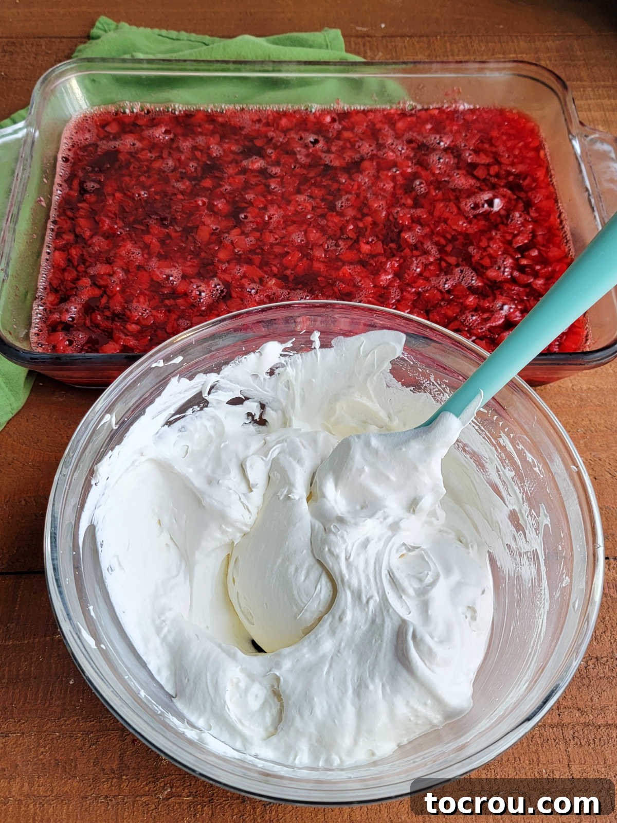 A bowl filled with a light and fluffy cream cheese and Cool Whip mixture, ready to be spread over a set pan of cranberry Jello salad in the background.