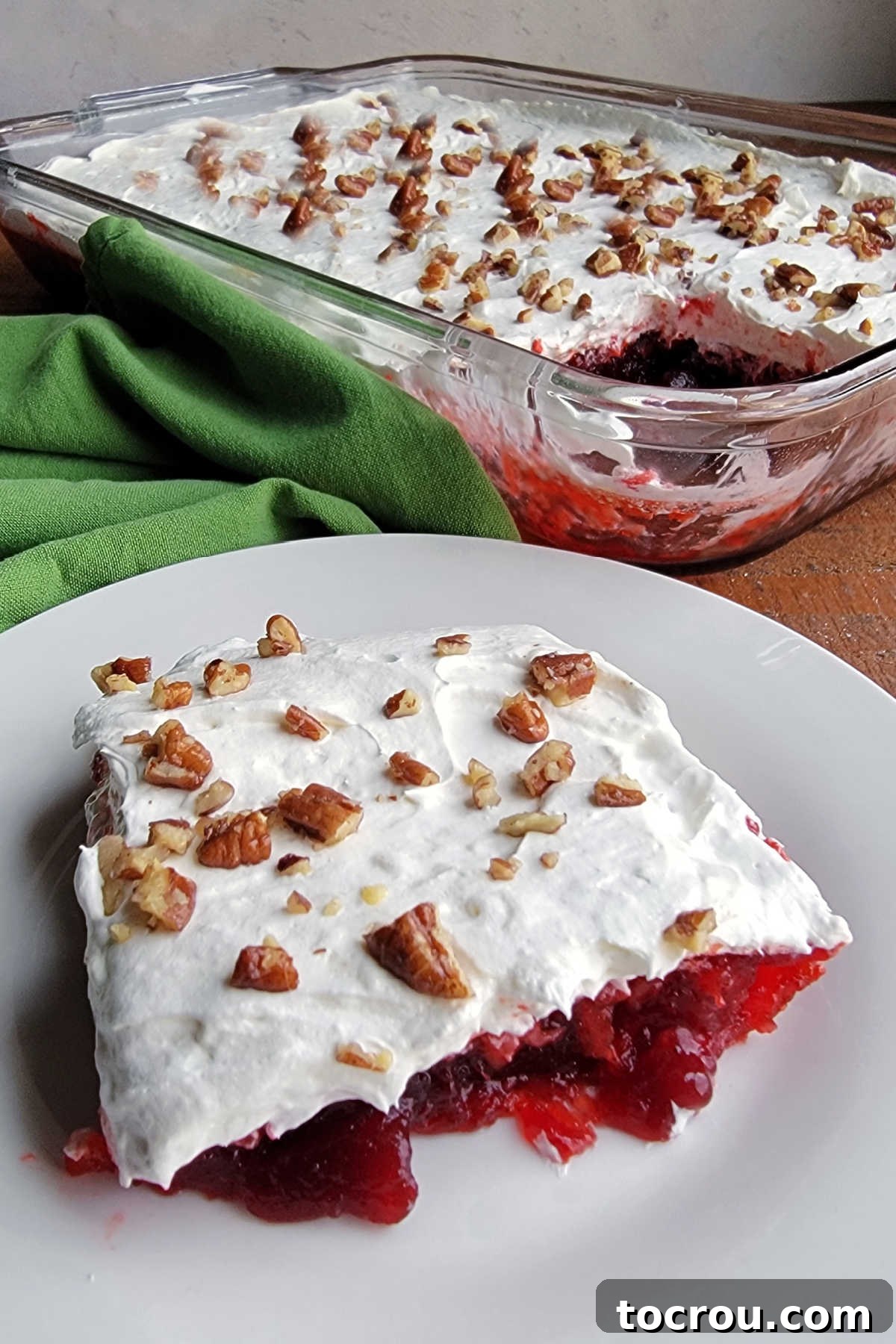 A perfect square of frosted cranberry Jello salad on a plate, with the remaining salad in a glass pan visible in the soft-focus background, ready to be served.