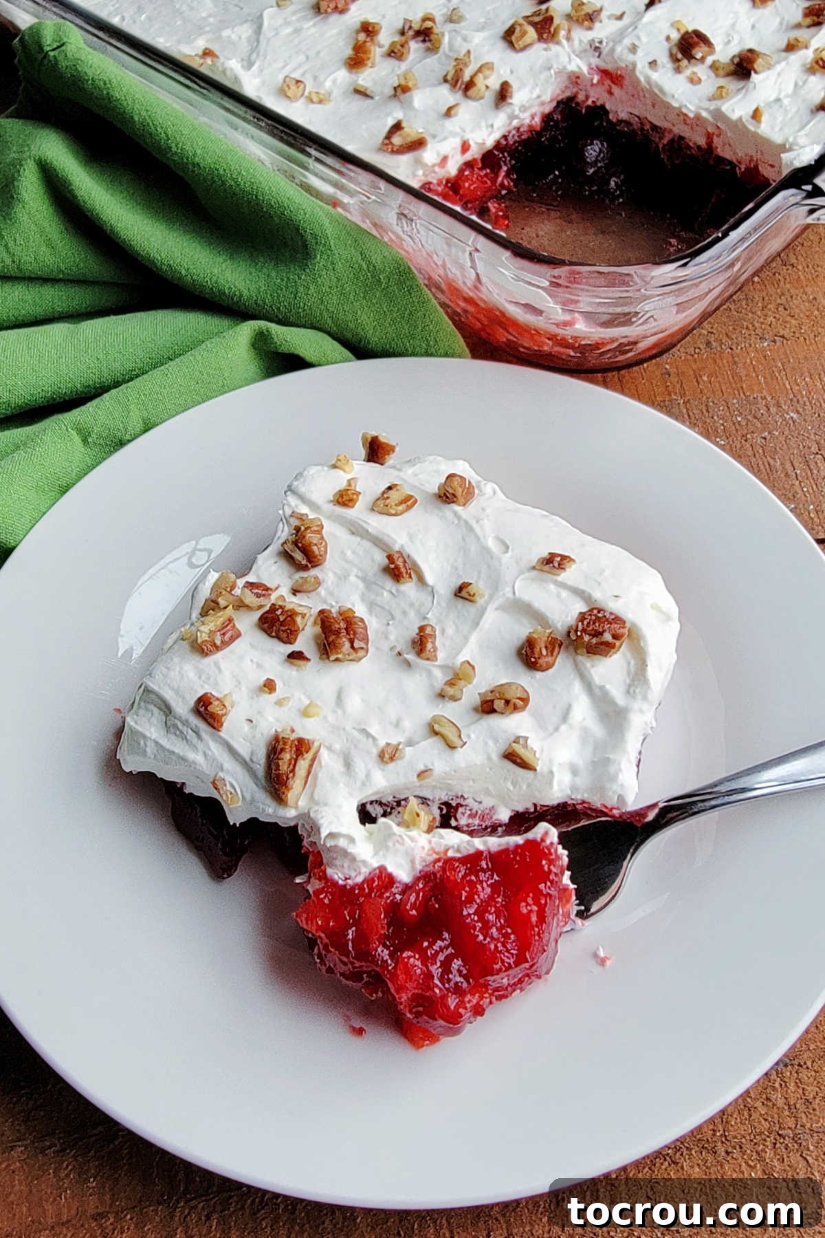 A fork holding a bite of frosted cranberry Jello salad, clearly showing the bright red Jello base with bits of pineapple, a generous layer of fluffy white cream cheese, and a sprinkle of toasted pecans.
