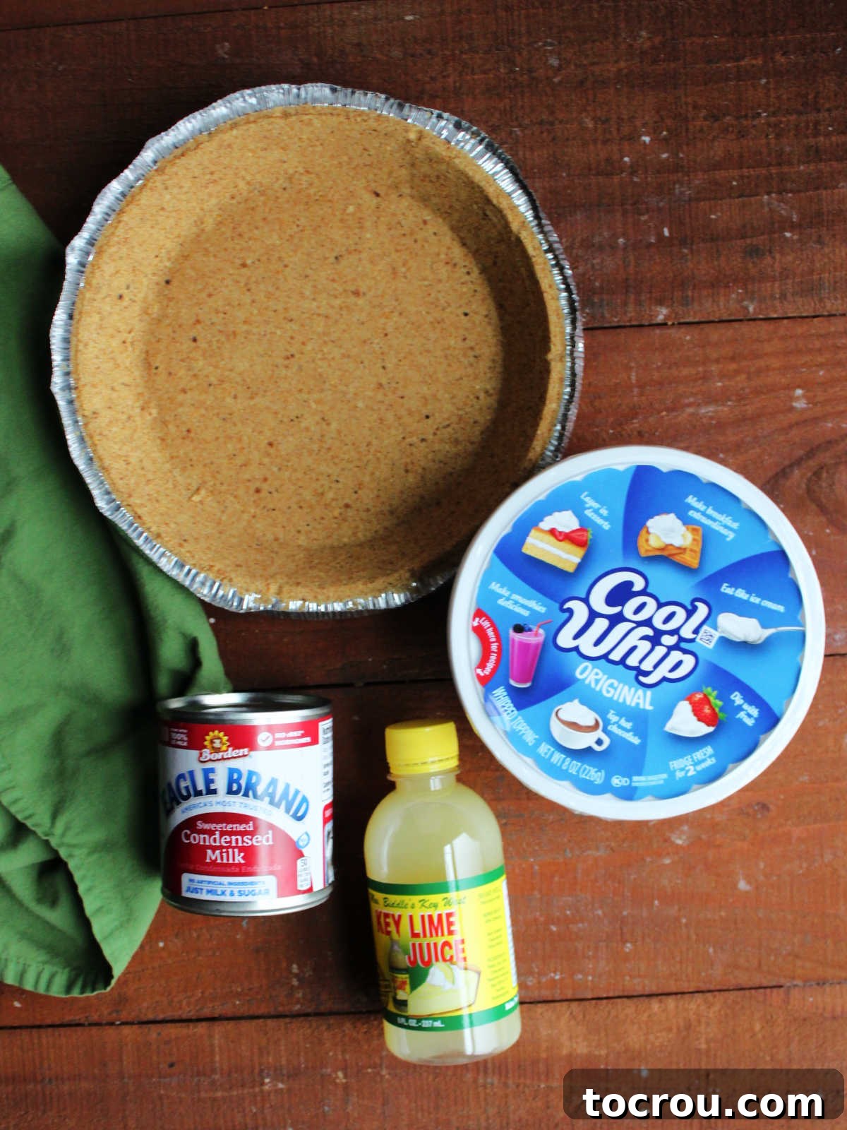 Key Lime Pie Ingredients Ingredients for no-bake key lime pie: a pre-made graham cracker crust, a tub of Cool Whip, a bottle of key lime juice, and a can of sweetened condensed milk, arranged on a kitchen counter.