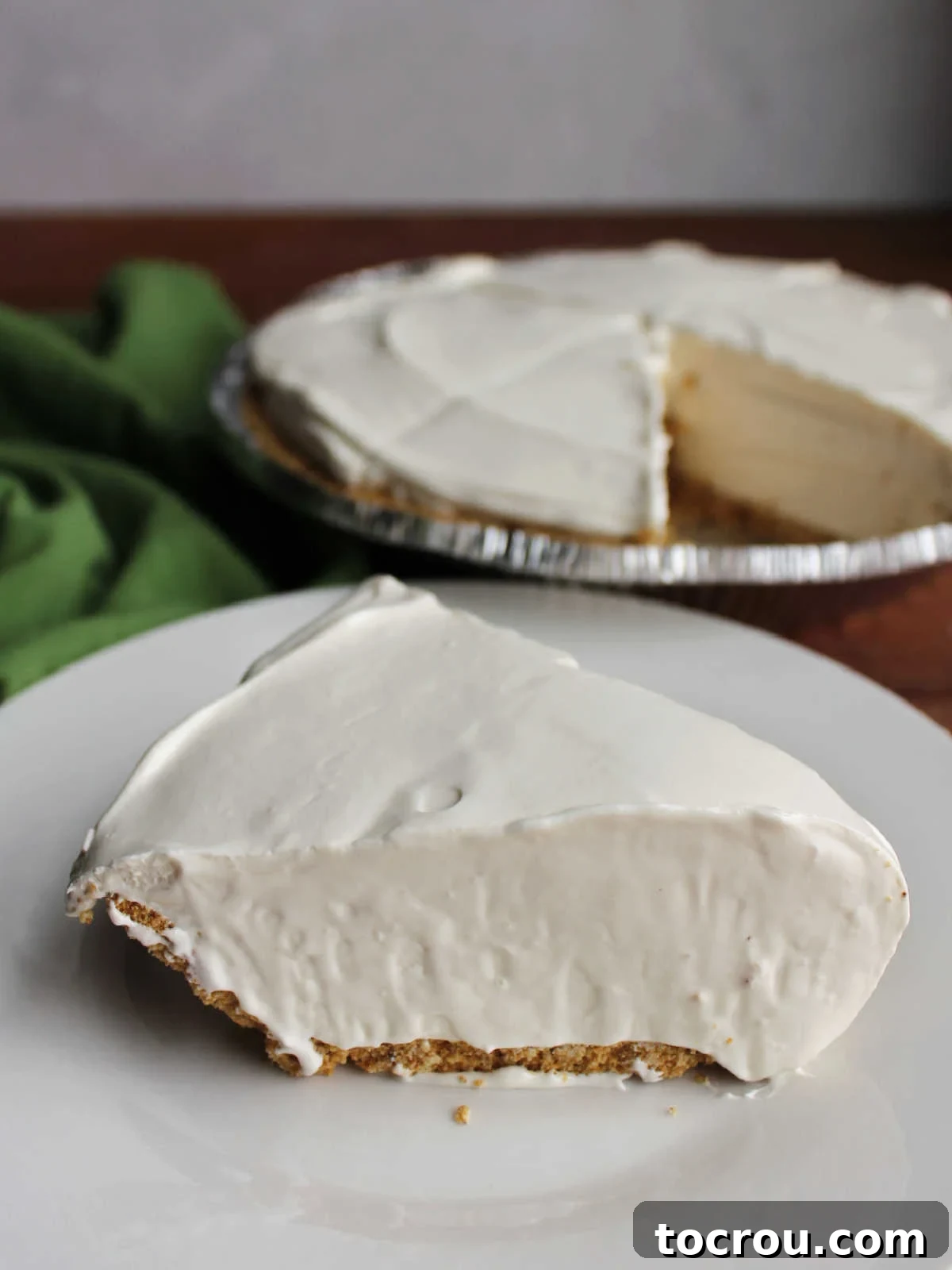 Individual Slice of Key Lime Pie A single slice of no-bake key lime pie presented on a small white plate, with the rest of the pie blurred in the background, highlighting the smooth texture and golden crust.