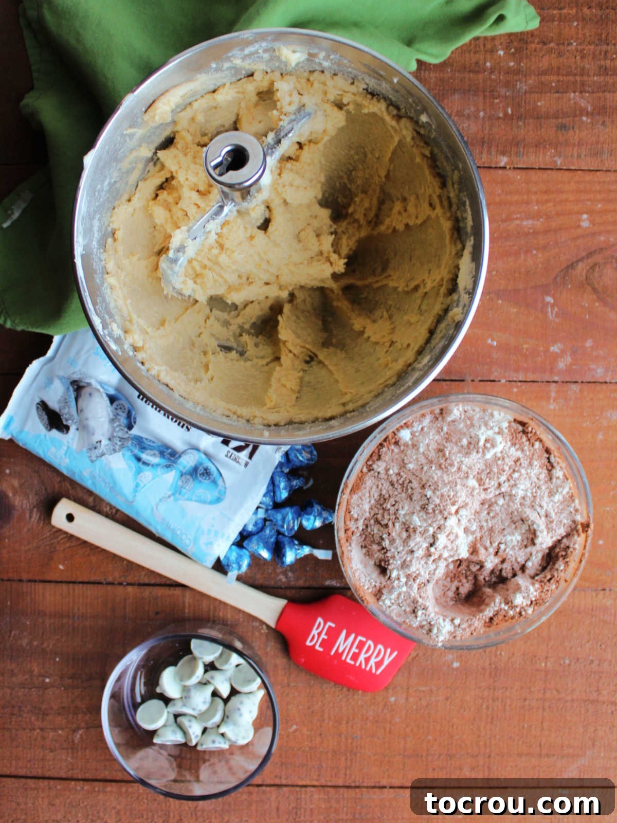 Oreo Dream Blossoms 3 A scene illustrating the cookie-making process: a mixer bowl contains a creamy butter, sugar, and egg mixture. Beside it, a separate bowl holds the dry ingredients—flour, cocoa powder, baking soda, and salt. In the foreground, a bowl of unwrapped Cookies and Cream Kisses anticipates their role in the finished treats.