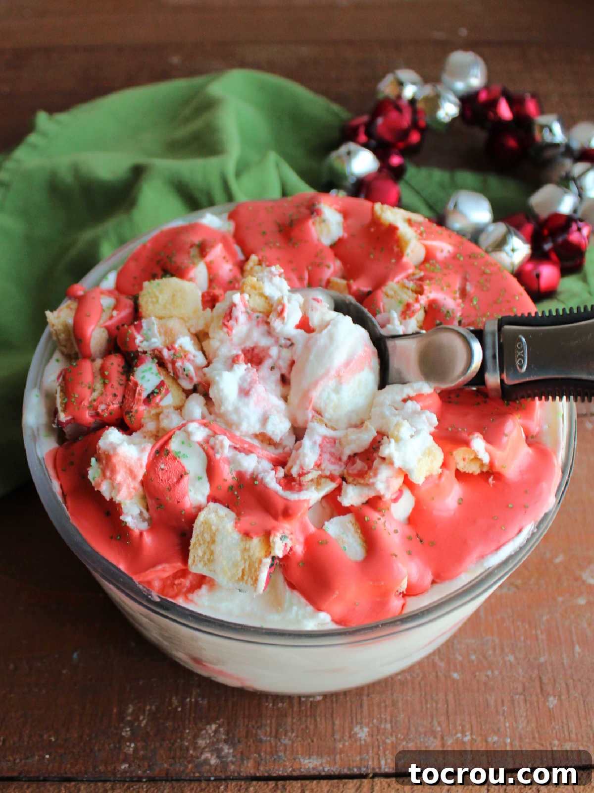 An ice cream scoop retrieving a generous portion of Christmas tree cake ice cream from a container, clearly showing the creamy vanilla base studded with substantial chunks of snack cake and a beautiful swirl of red buttercream within.