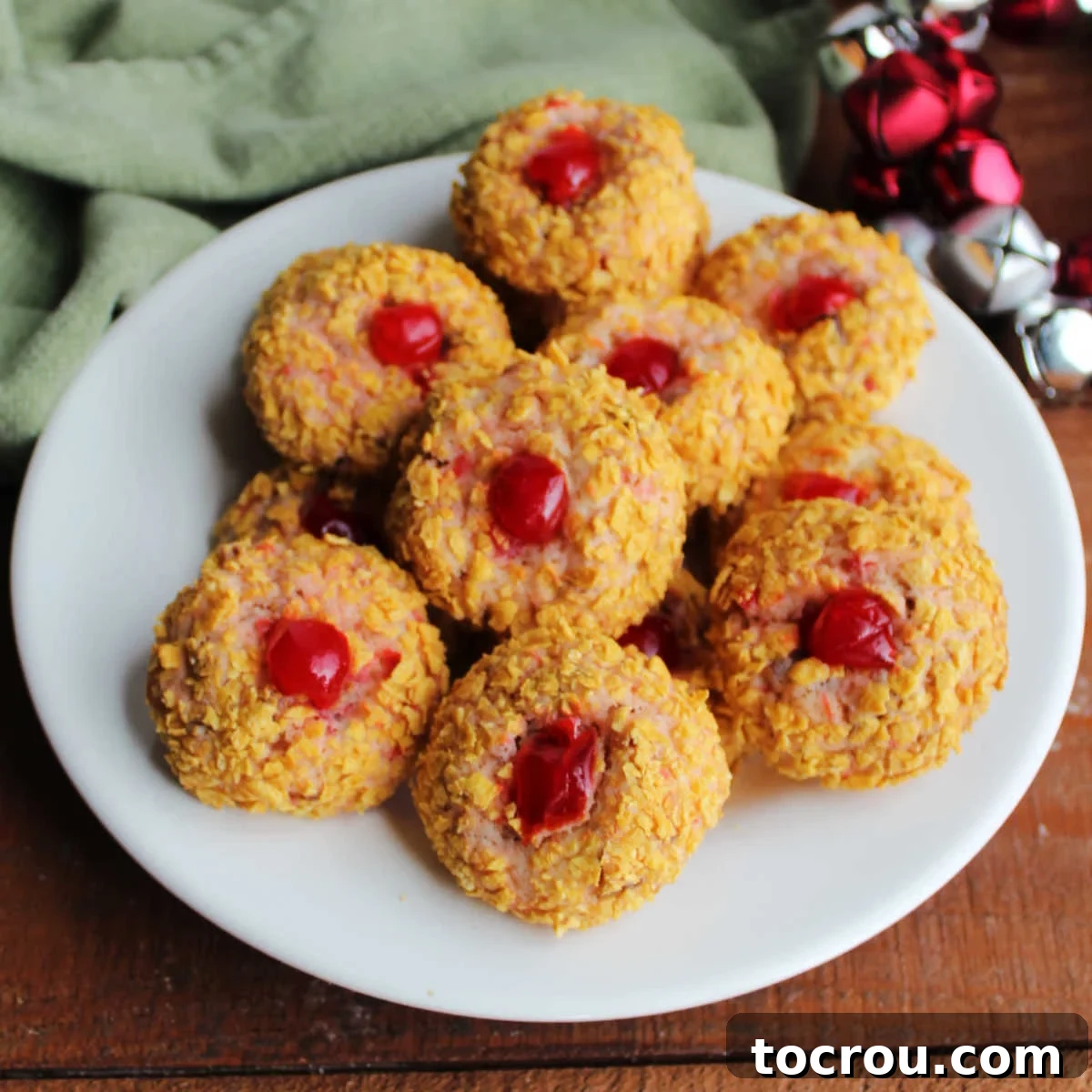 Plate piled with cherry winks cookies with light pink cookie showing through cornflake crumb exterior with a piece of a bright red maraschino cherry in the middle.